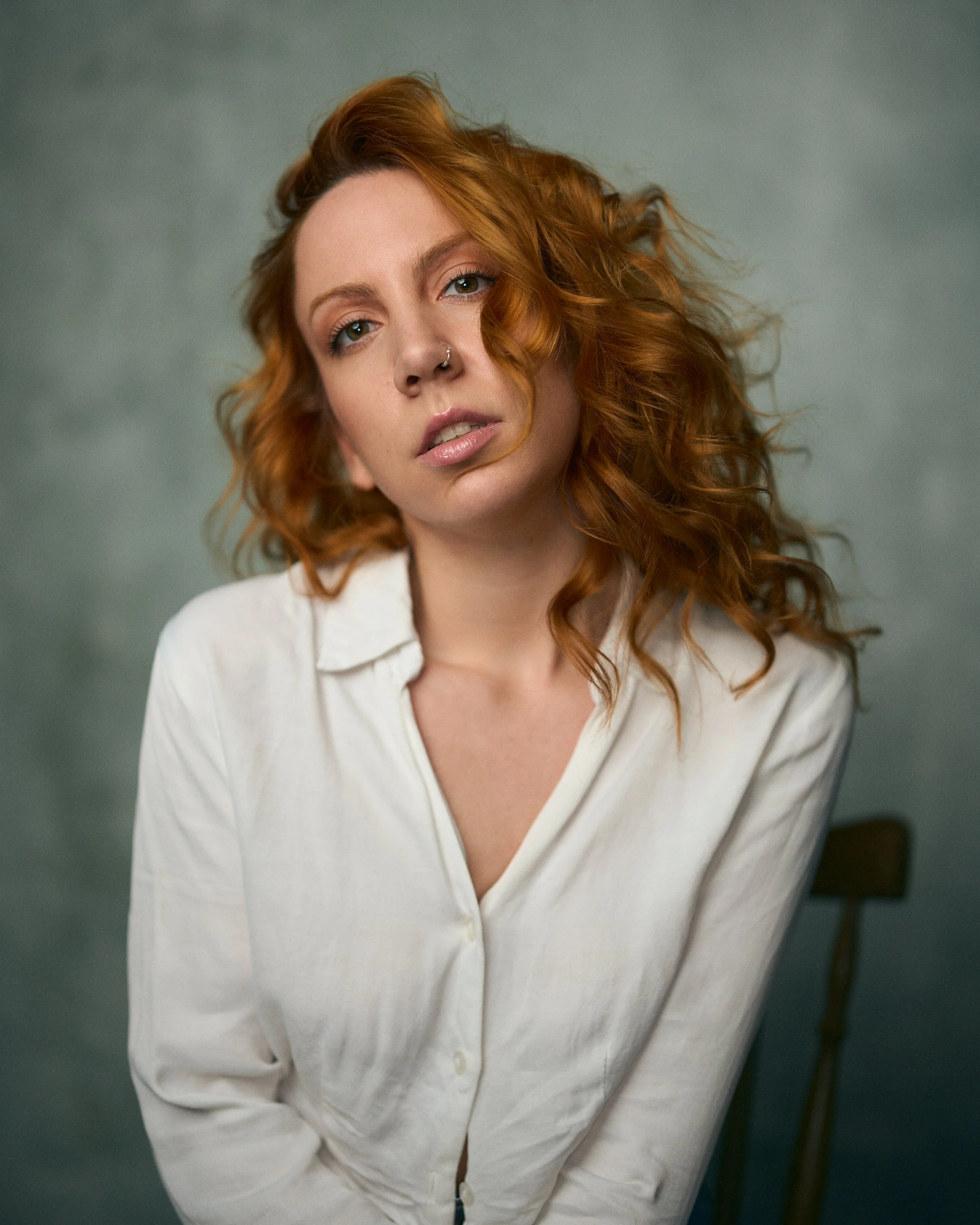Portrait of a woman with curly red hair, wearing a white button-down shirt, looking into the camera with a neutral expression, seated on a wooden chair against a gray background.