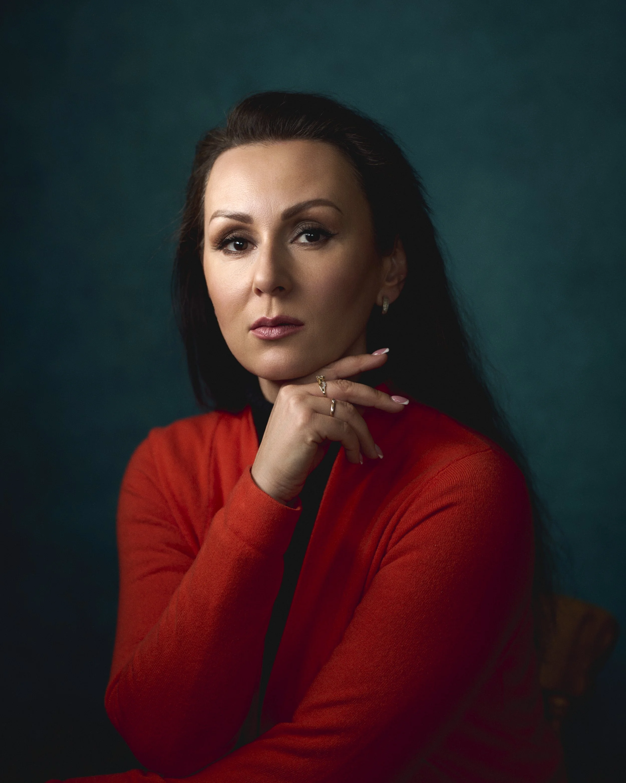 Portrait of a woman with dark hair wearing a red blazer, posing with her hand near her chin, against a dark teal background.