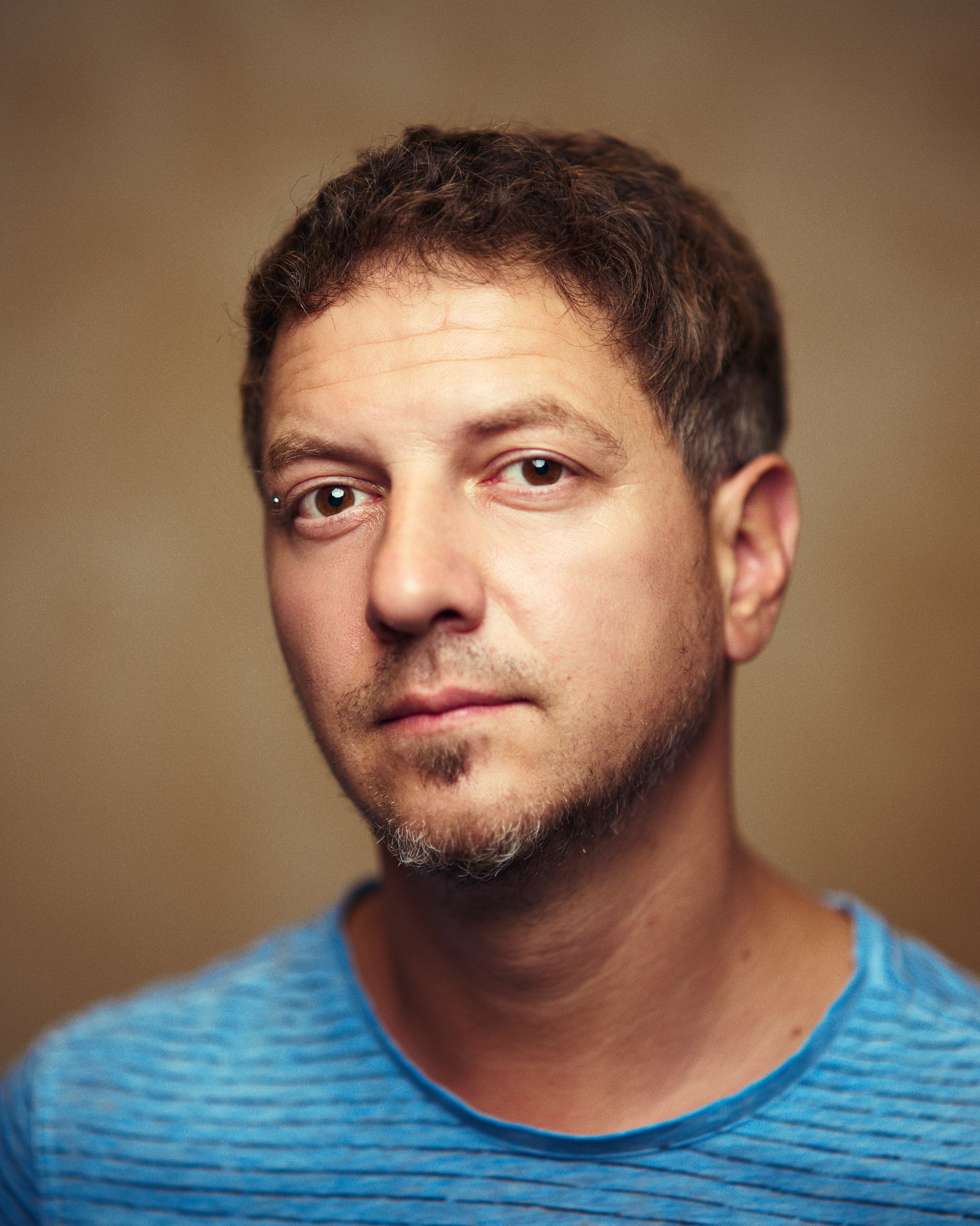 A man with short brown hair and a small goatee, wearing a blue and black striped shirt, looking at the camera with a neutral expression.
