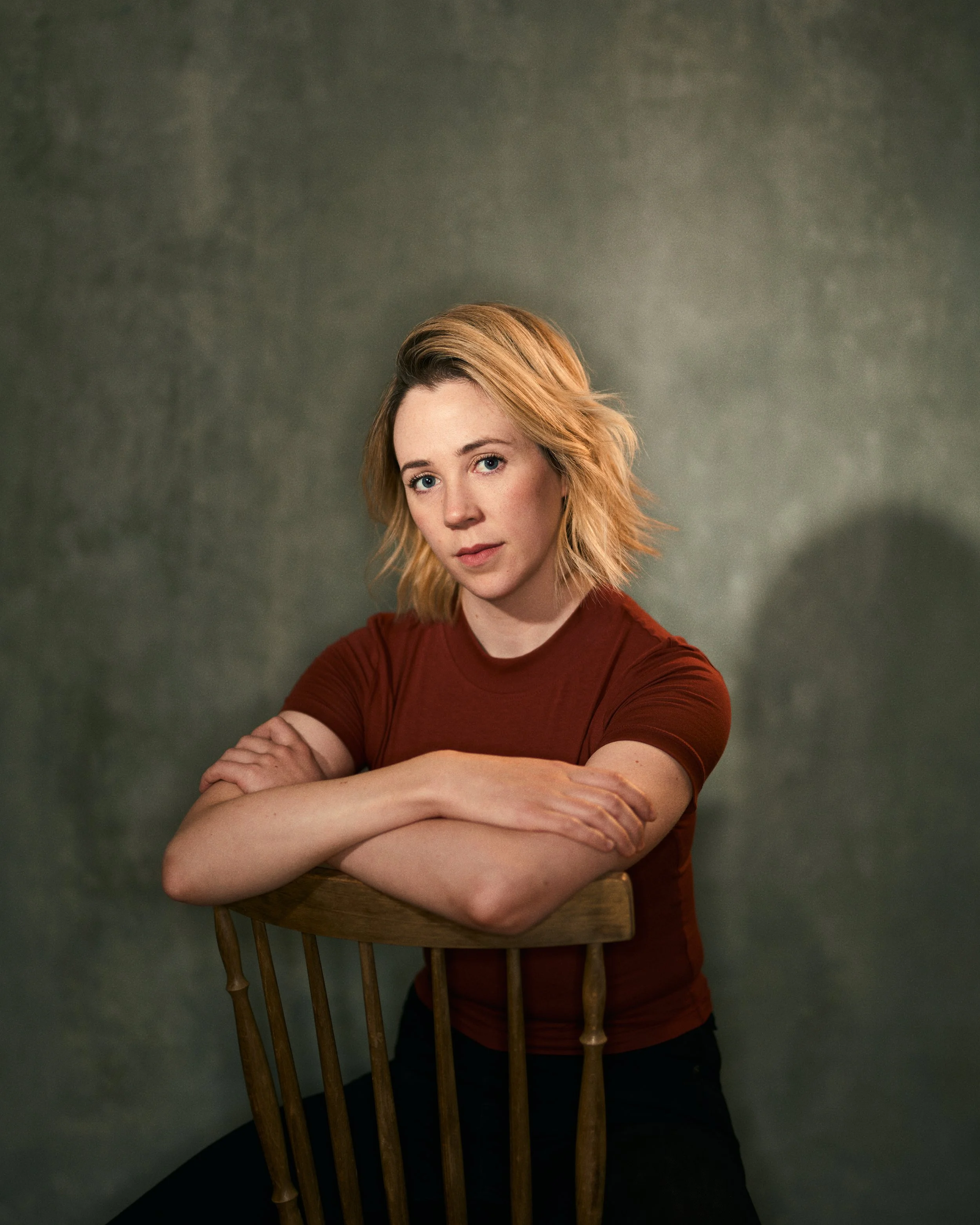 A woman with shoulder-length blonde hair and fair skin, wearing a red t-shirt, sitting on a wooden chair with her arms crossed, looking directly at the camera against a plain gray background.