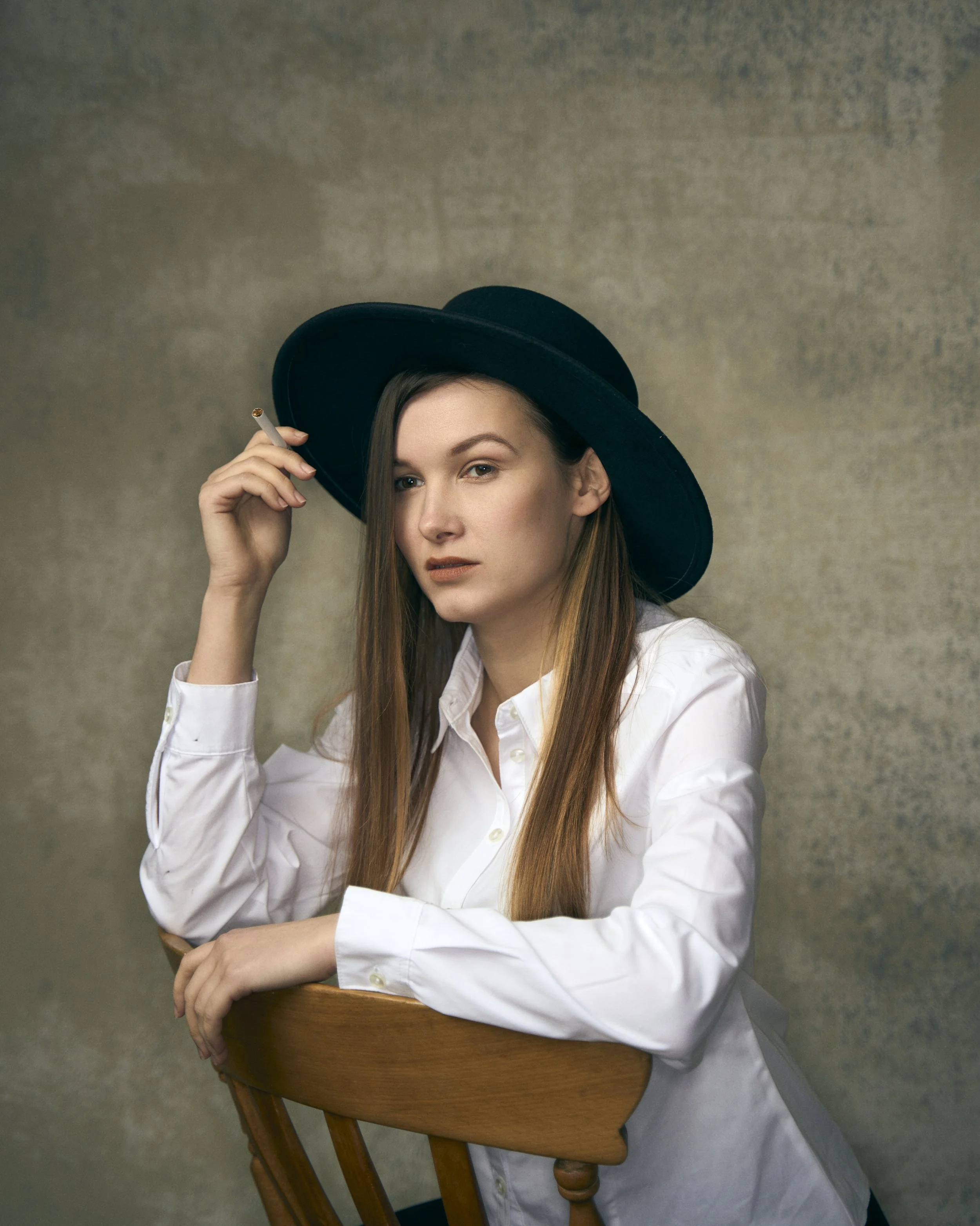 A young woman wearing a white shirt, black wide-brimmed hat, and holding a cigarette, sitting on a wooden chair against a textured beige background.