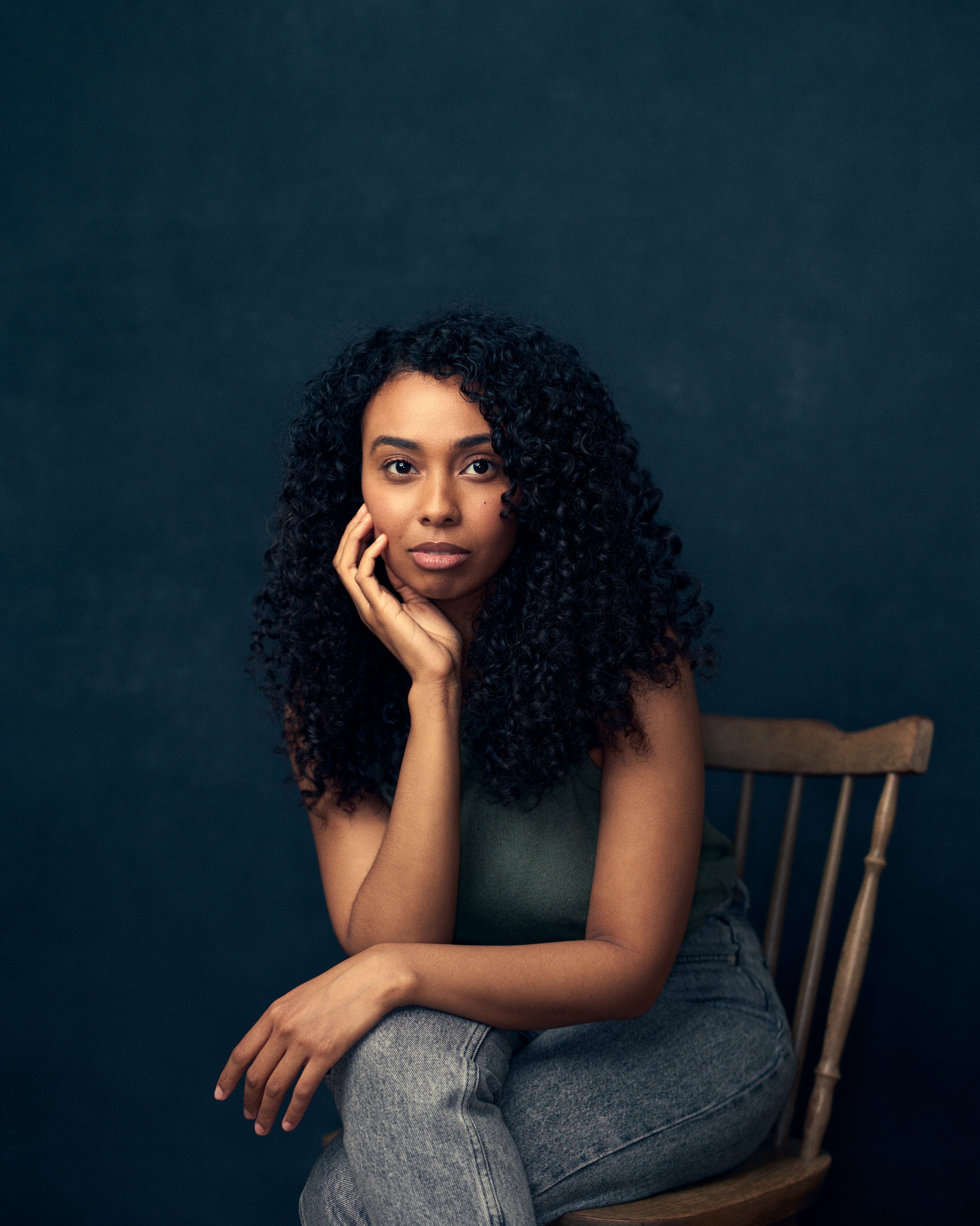 Portrait of a woman with curly black hair sitting on a wooden chair against a dark background, resting her chin on her hand and looking at the camera.