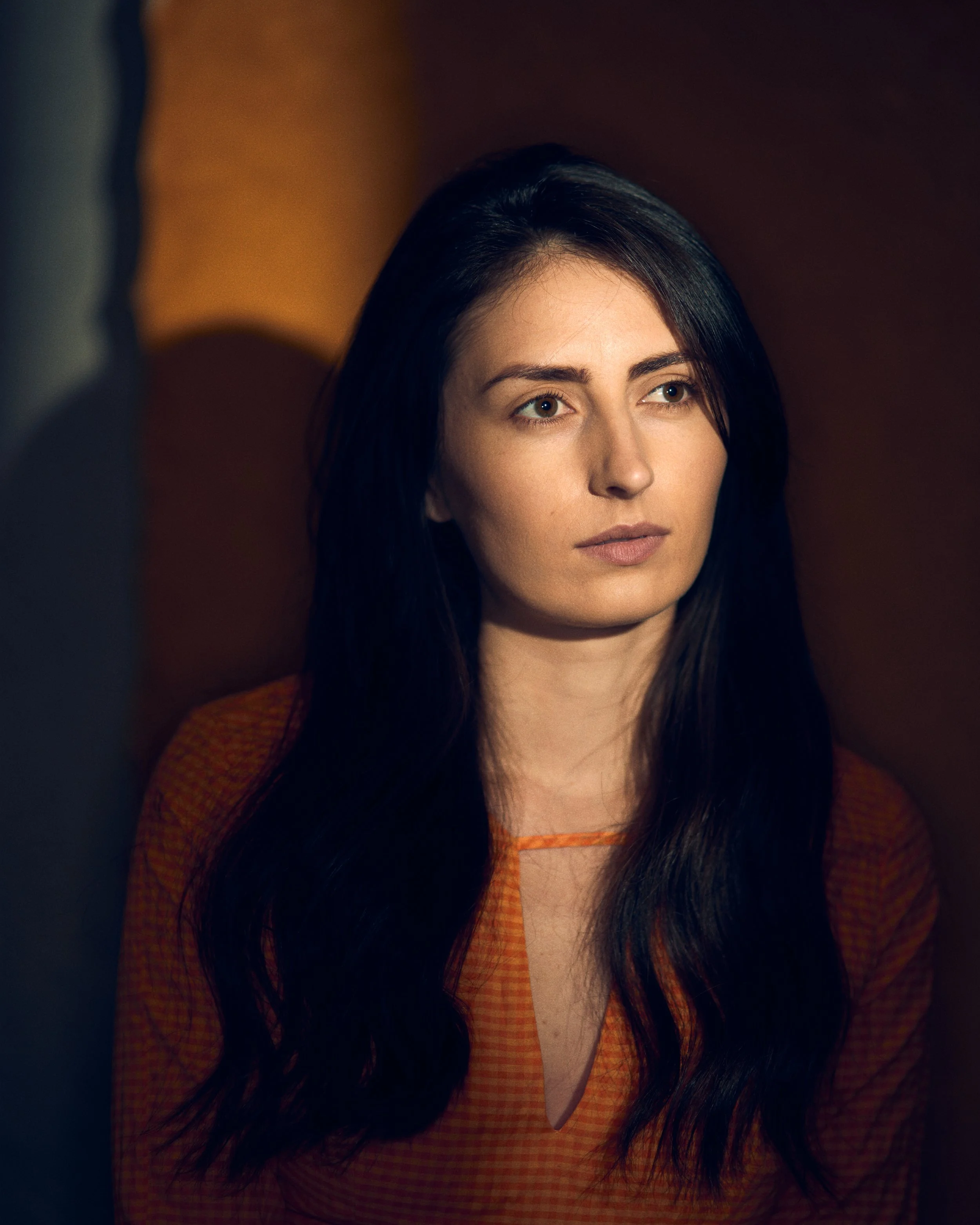 Close-up of a woman with long dark hair, sitting indoors, wearing an orange top, looking pensively to the side with a neutral expression.