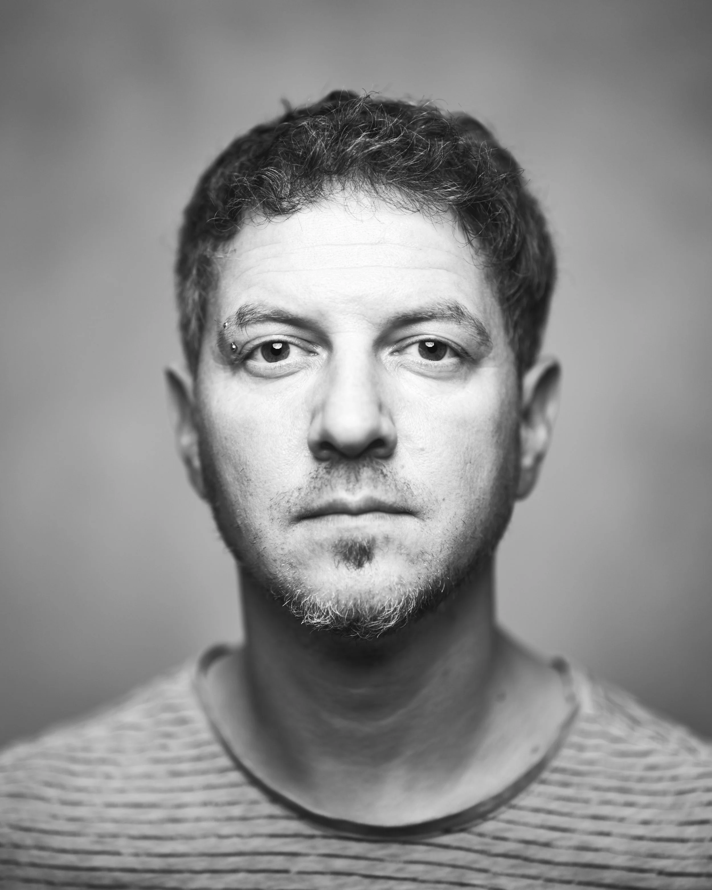 Black and white portrait of a man with short, curly hair, a small beard, and a facial piercing above the left eyebrow, wearing a striped shirt.