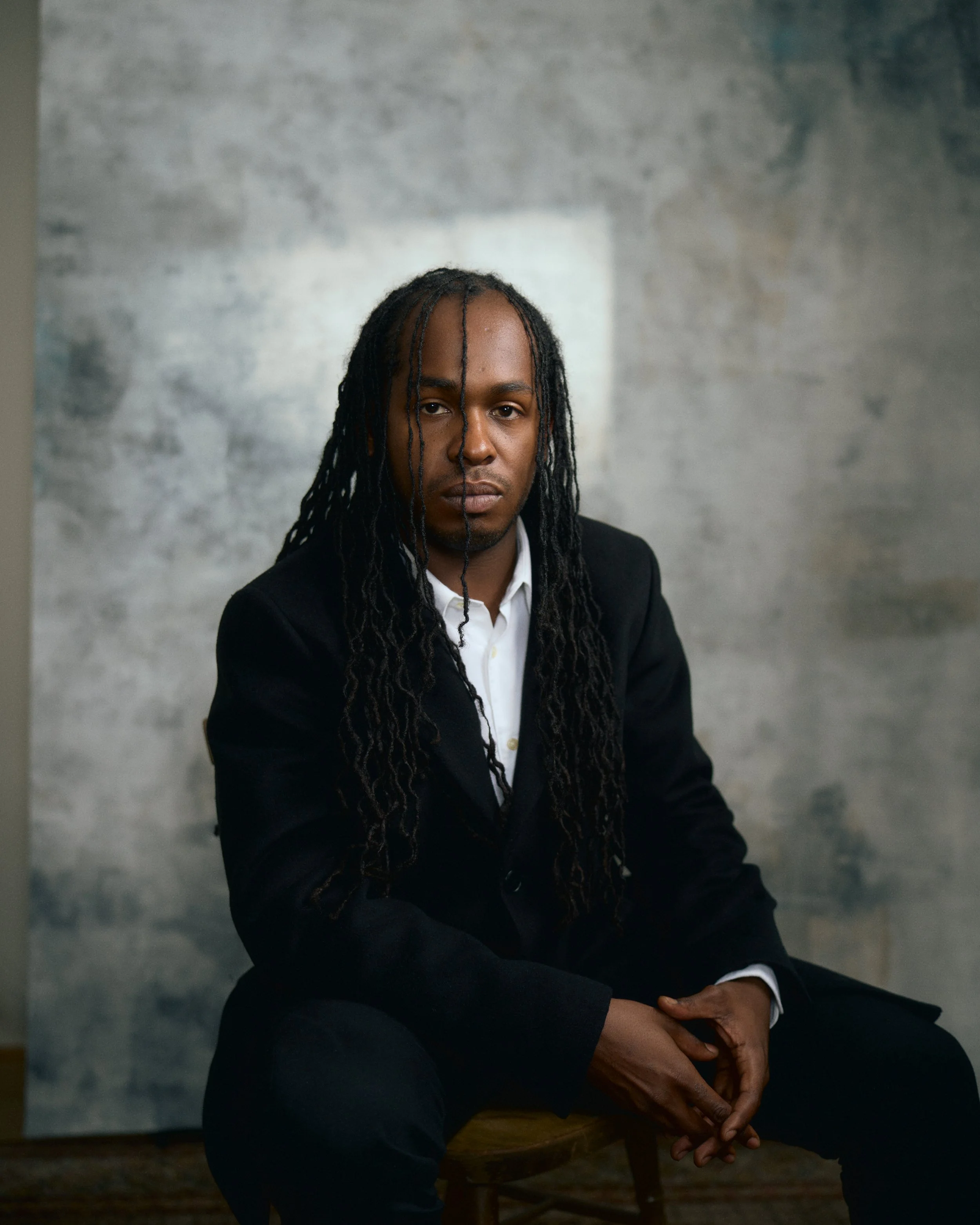 A man with long dreadlocks wearing a black suit and white shirt sitting on a wooden chair in front of a textured grey wall.