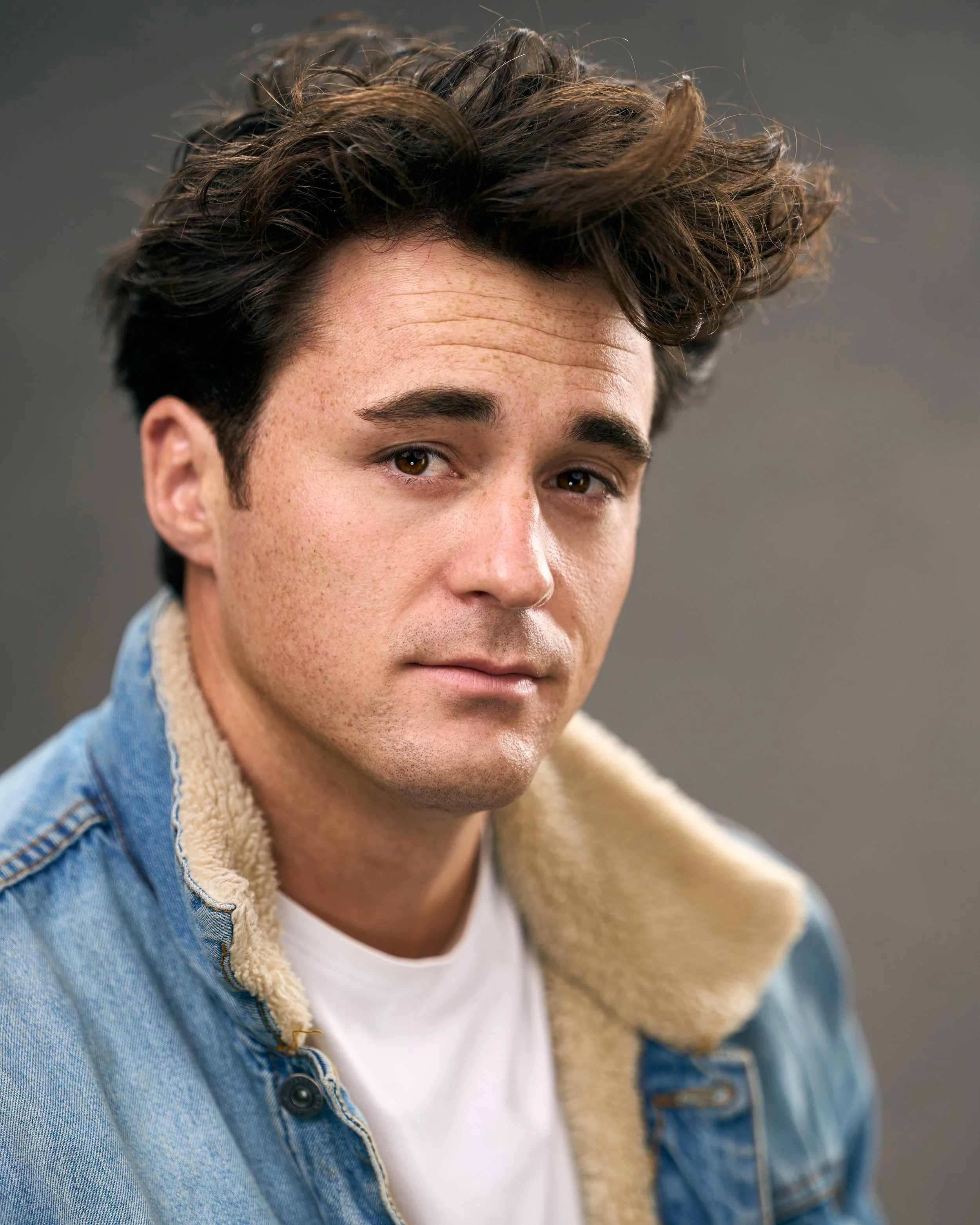 Close-up of a young man with brown, tousled hair, wearing a denim jacket with a shearling collar, and a white t-shirt, looking at the camera.