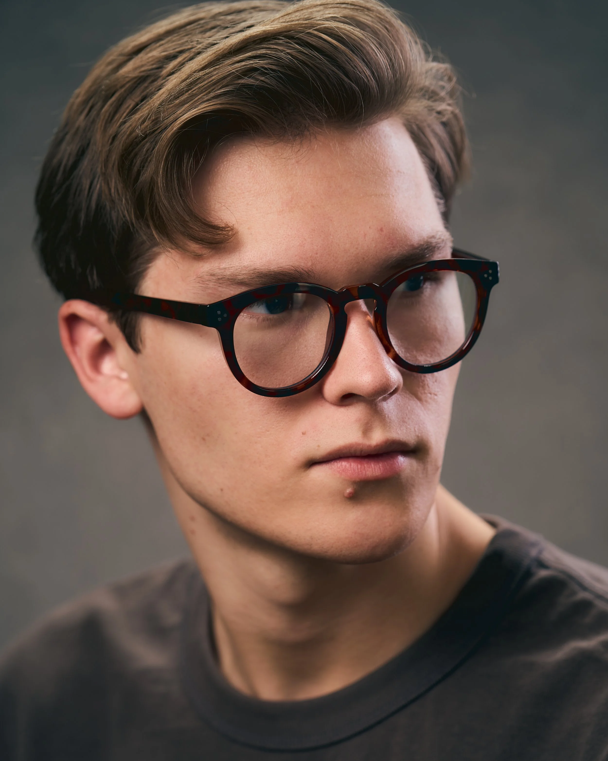 Close-up of a young man with brown hair, wearing large tortoiseshell glasses, and a dark T-shirt, looking to the side with a neutral expression.