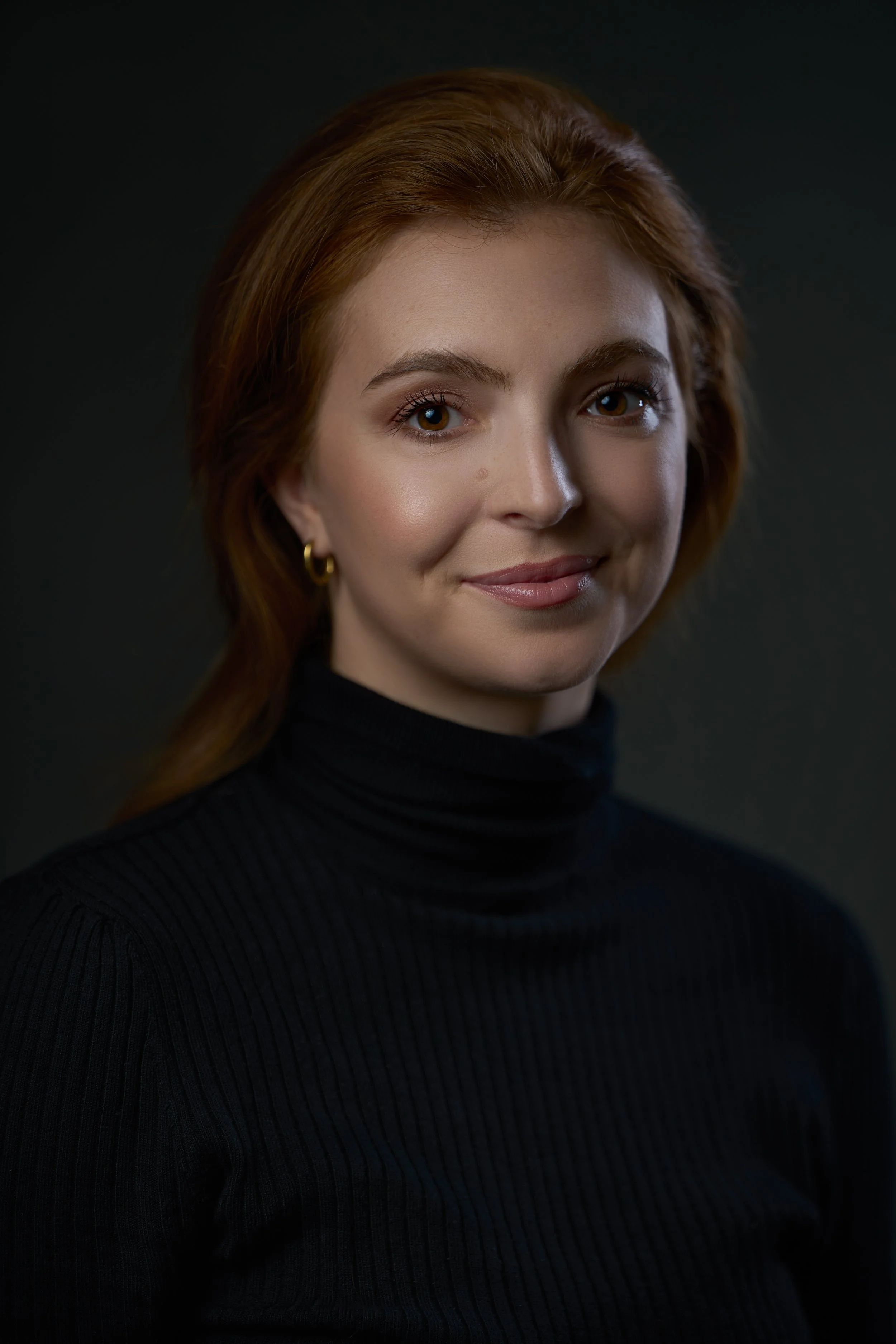 Portrait of a woman with red hair wearing a black turtleneck against a dark background.