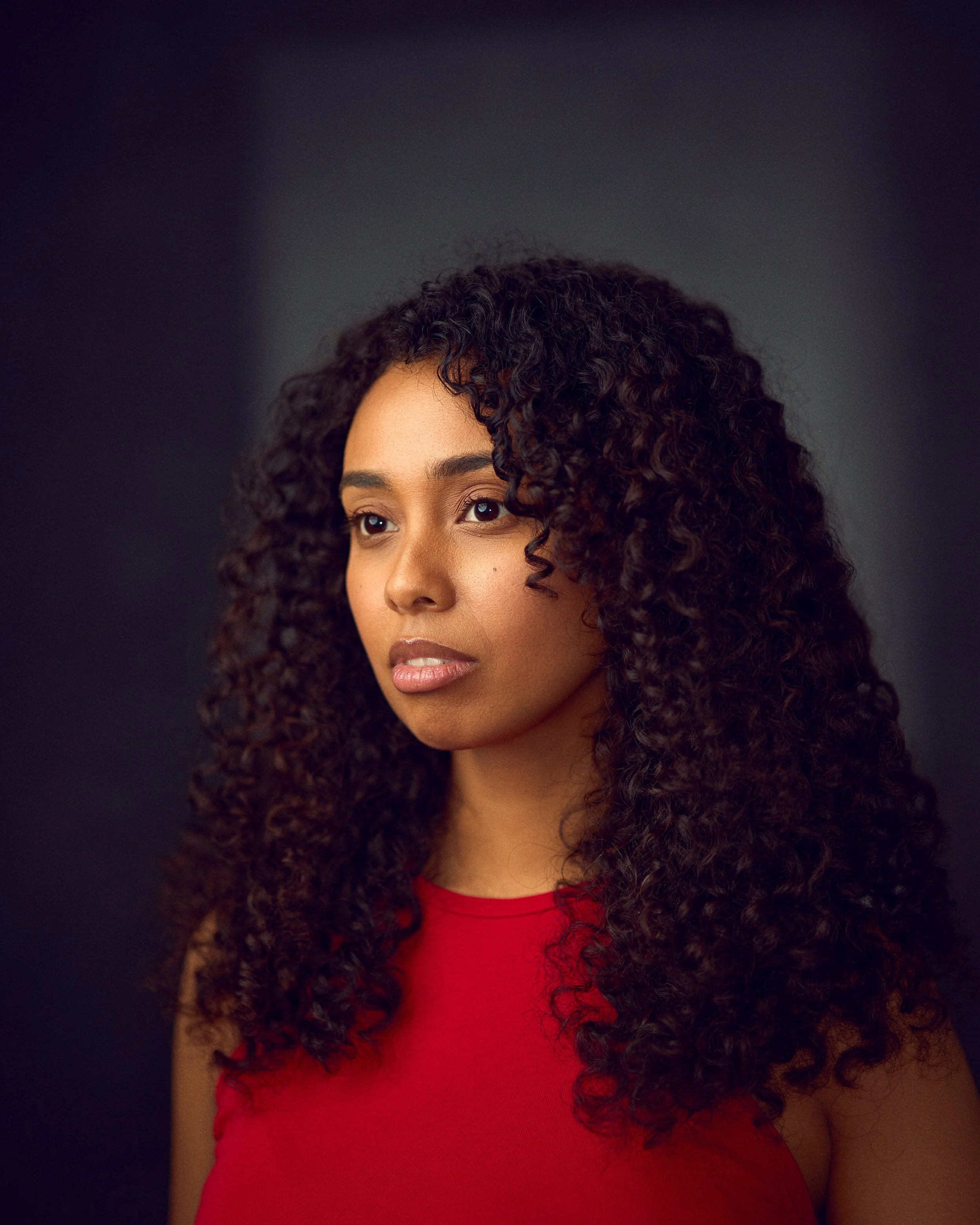 Portrait of a woman with curly dark hair wearing a red top against a dark background.