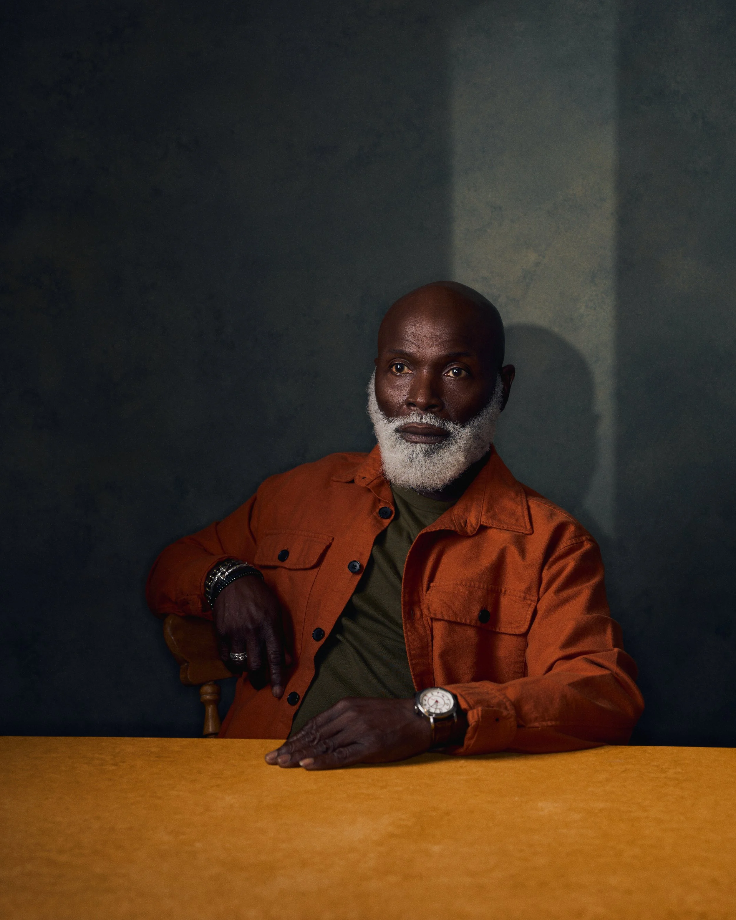 Portrait of a distinguished older Black man with a gray beard, wearing an orange jacket, sitting with his hands on a yellow table against a dark background.