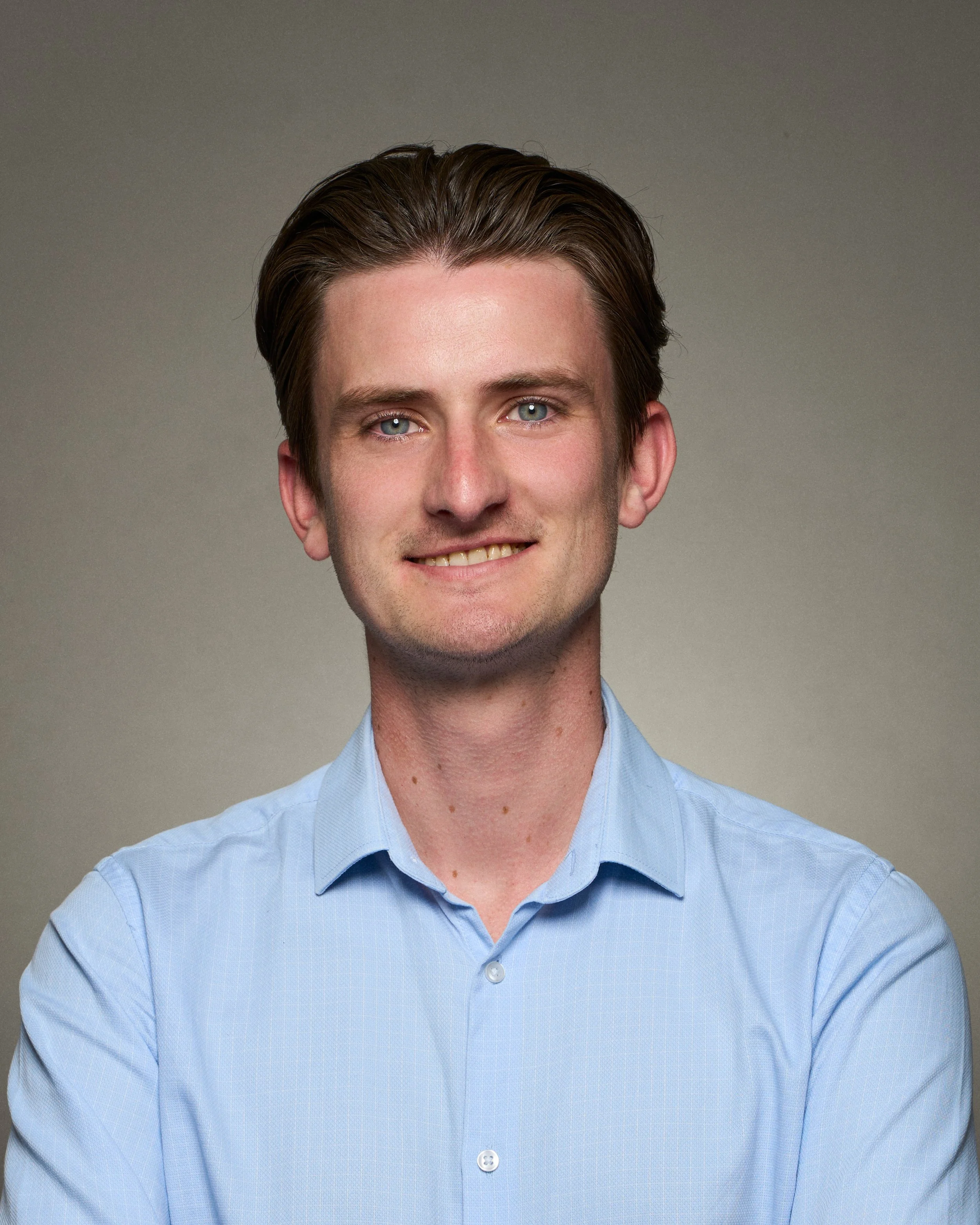 A young man with brown hair and blue eyes wearing a light blue button-down shirt, smiling, with a neutral background.