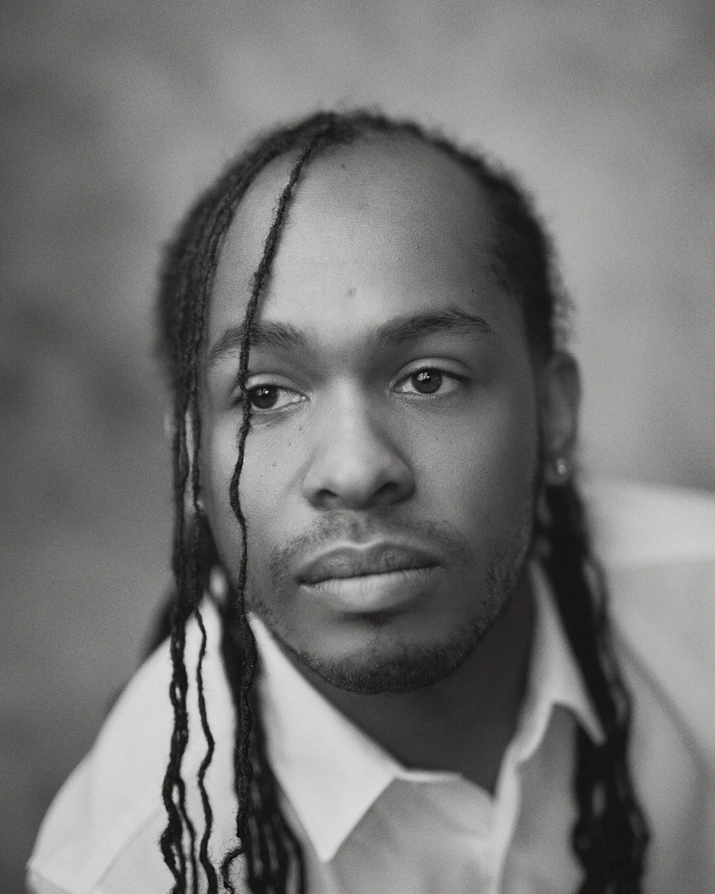Black and white portrait of a man with long braided hair, wearing a collared shirt, looking away from the camera.