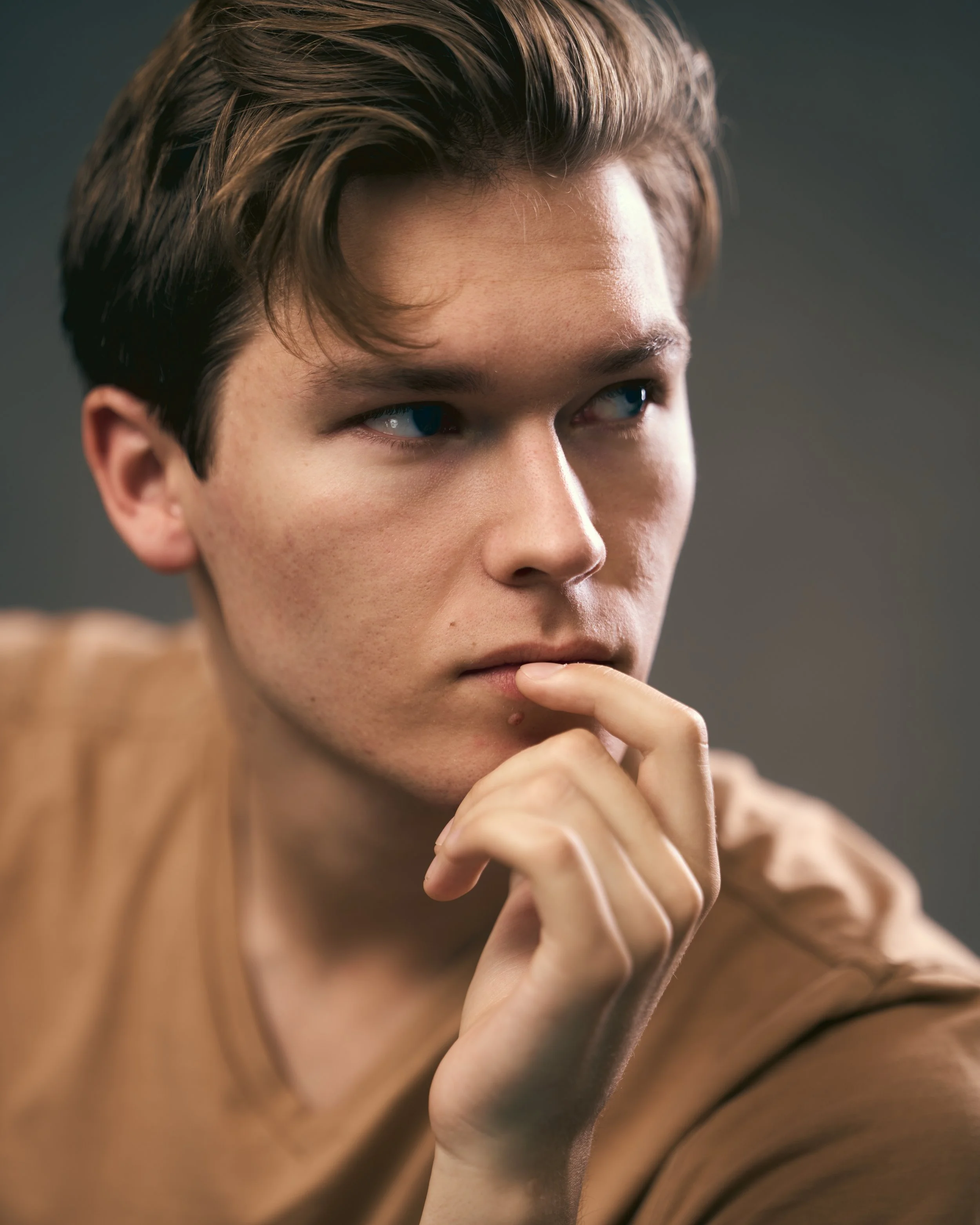 A young man with light skin and brown hair, thinking with his right hand touching his lips, wearing a tan shirt against a dark blurred background.