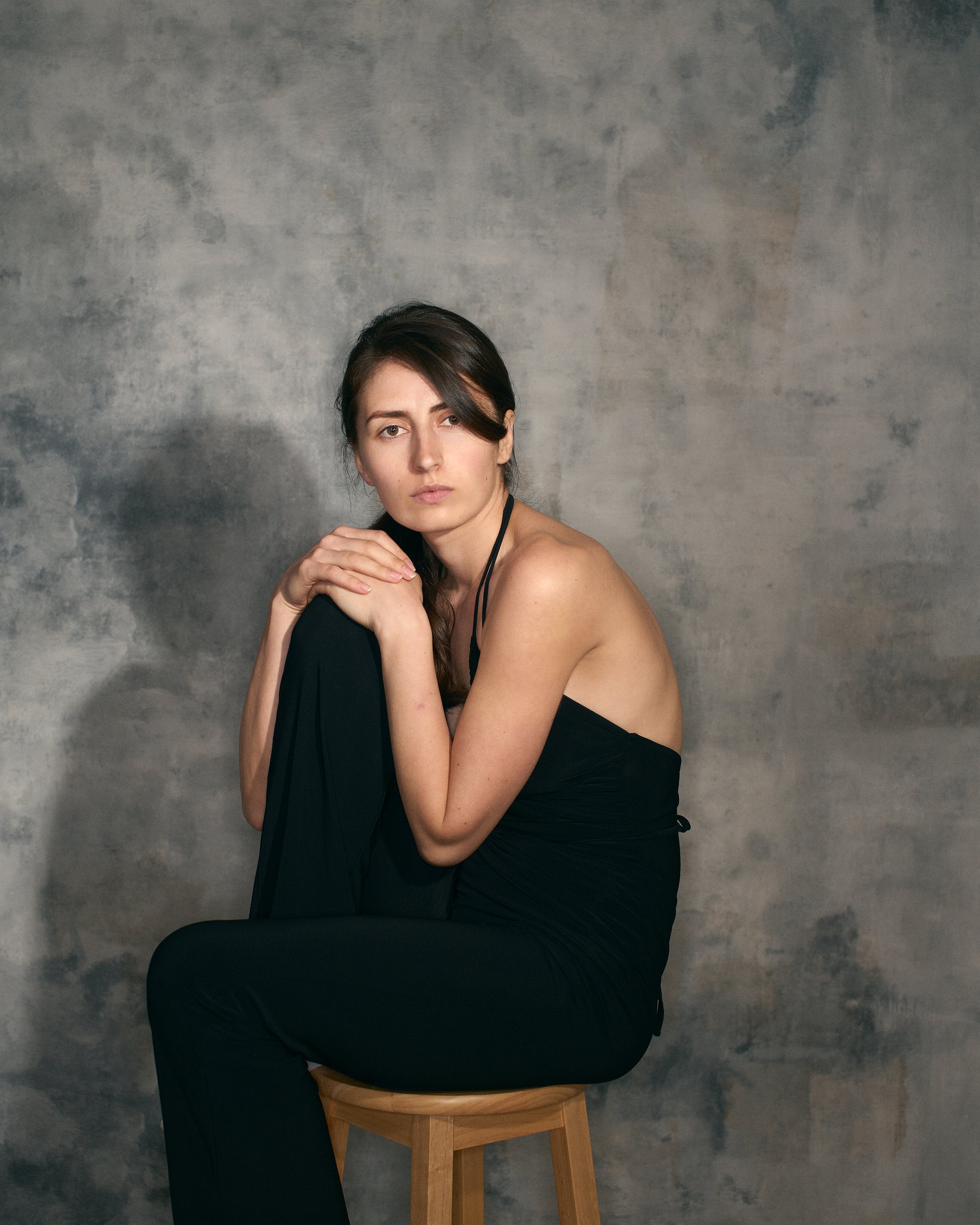 A woman sitting on a wooden stool against a textured grey background, wearing a black strapless dress and holding a black garment, looking at the camera with a serious expression.