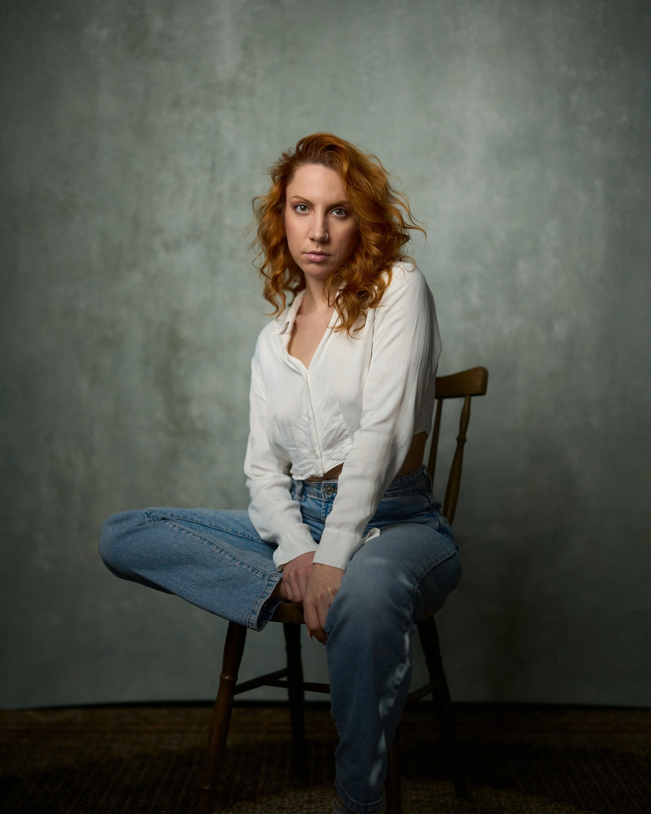 A young woman with curly red hair, wearing a white shirt and blue jeans, sits on a wooden chair with one leg crossed over the other, against a plain gray background.