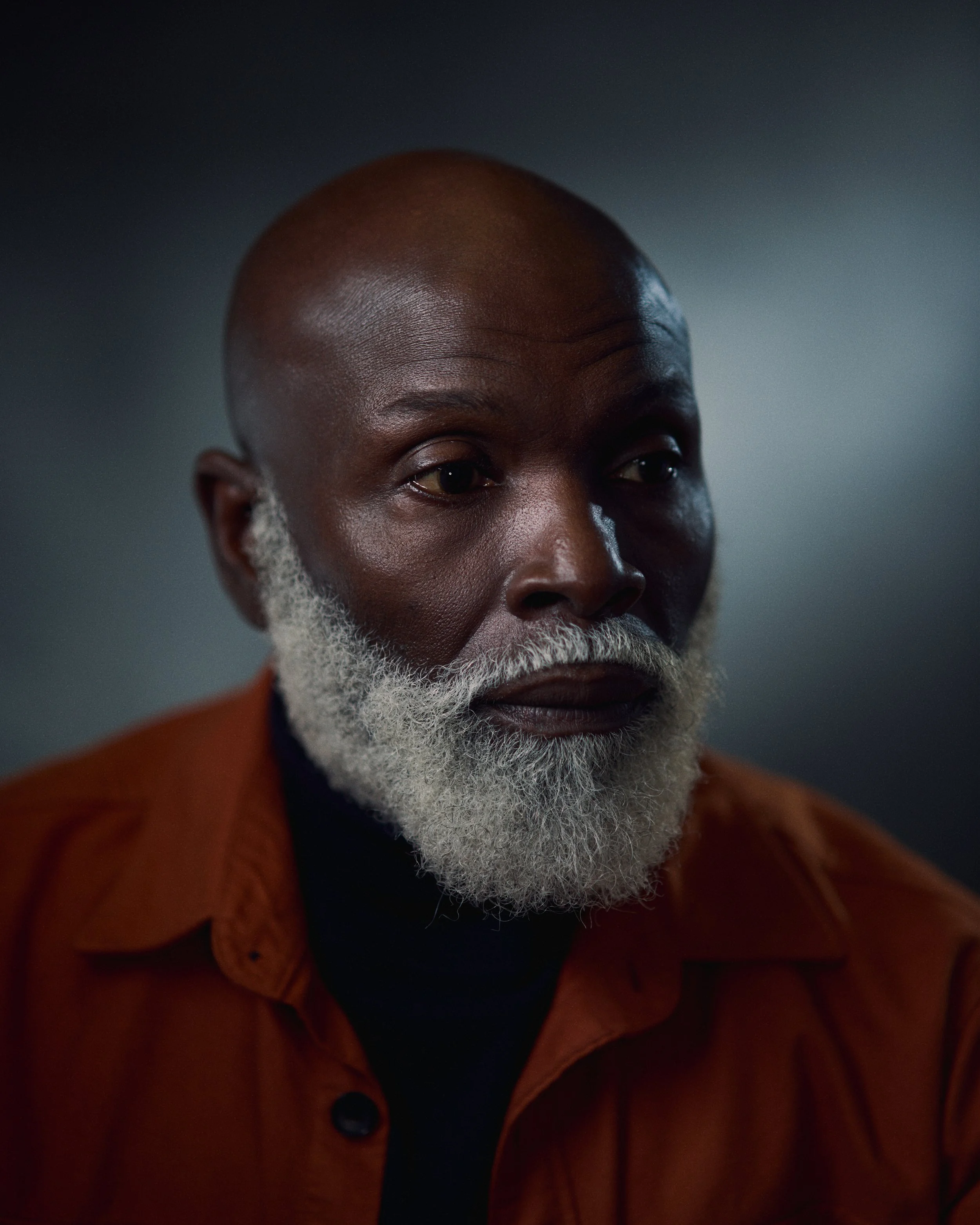 Portrait of an older Black man with a bald head and a white beard, wearing an orange shirt, against a dark background.