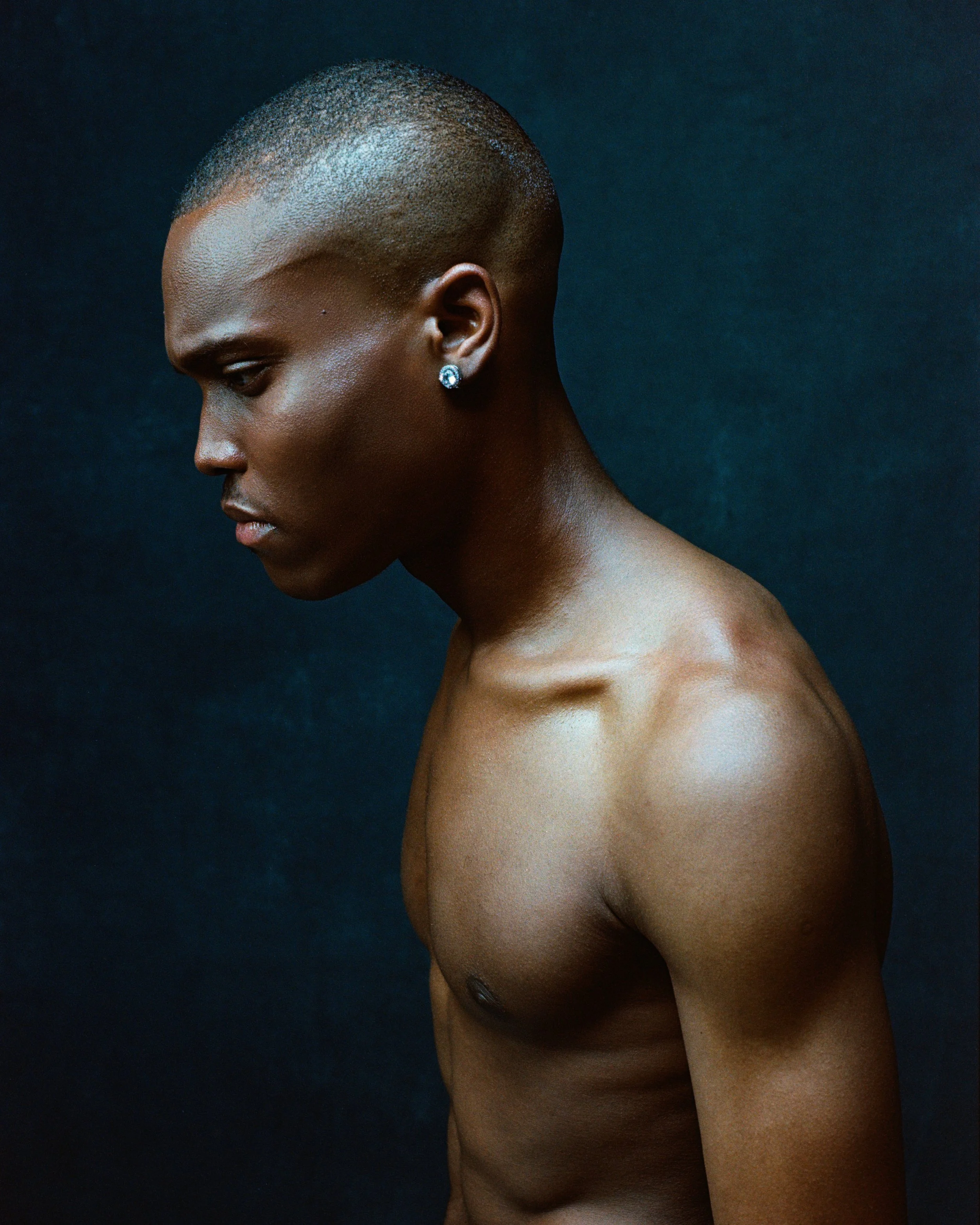 A side profile of a topless Black woman with a close-cropped haircut, wearing a diamond earring, against a dark textured background.