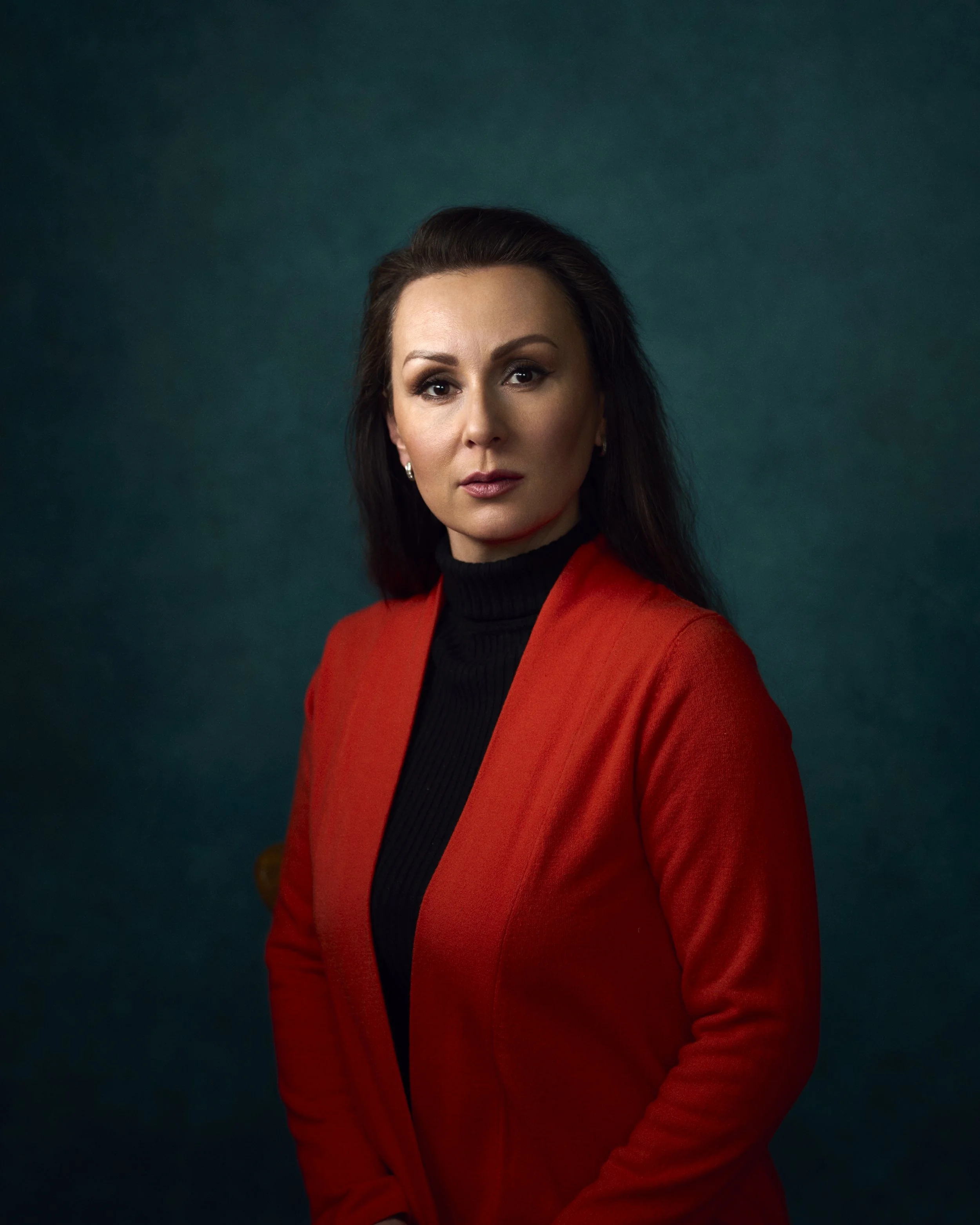 A woman with dark hair wearing a red blazer and black turtleneck posing against a dark teal background.