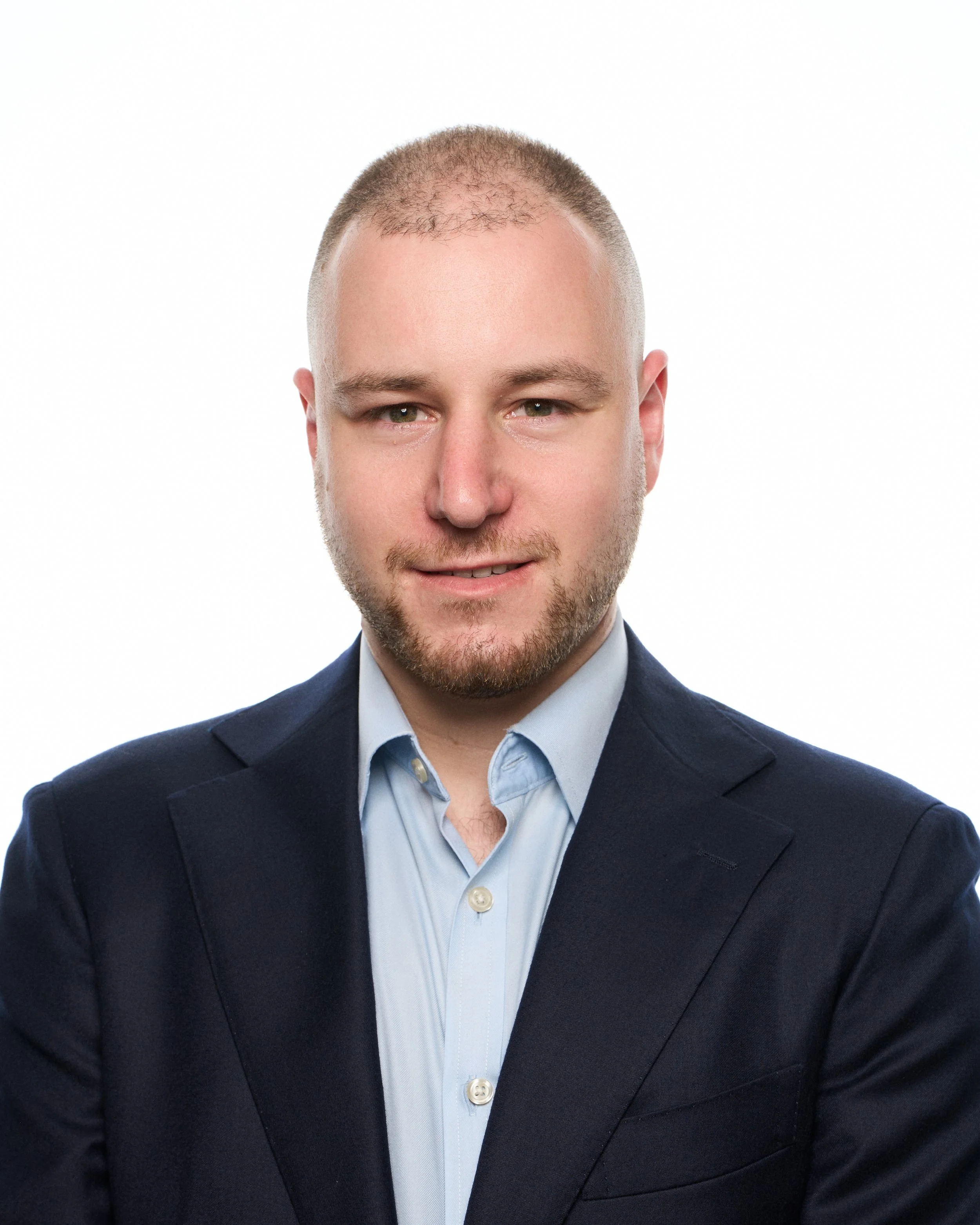 Headshot of a young man with light skin, short hair, and a beard, wearing a dark suit jacket and light blue collared shirt, against a white background.