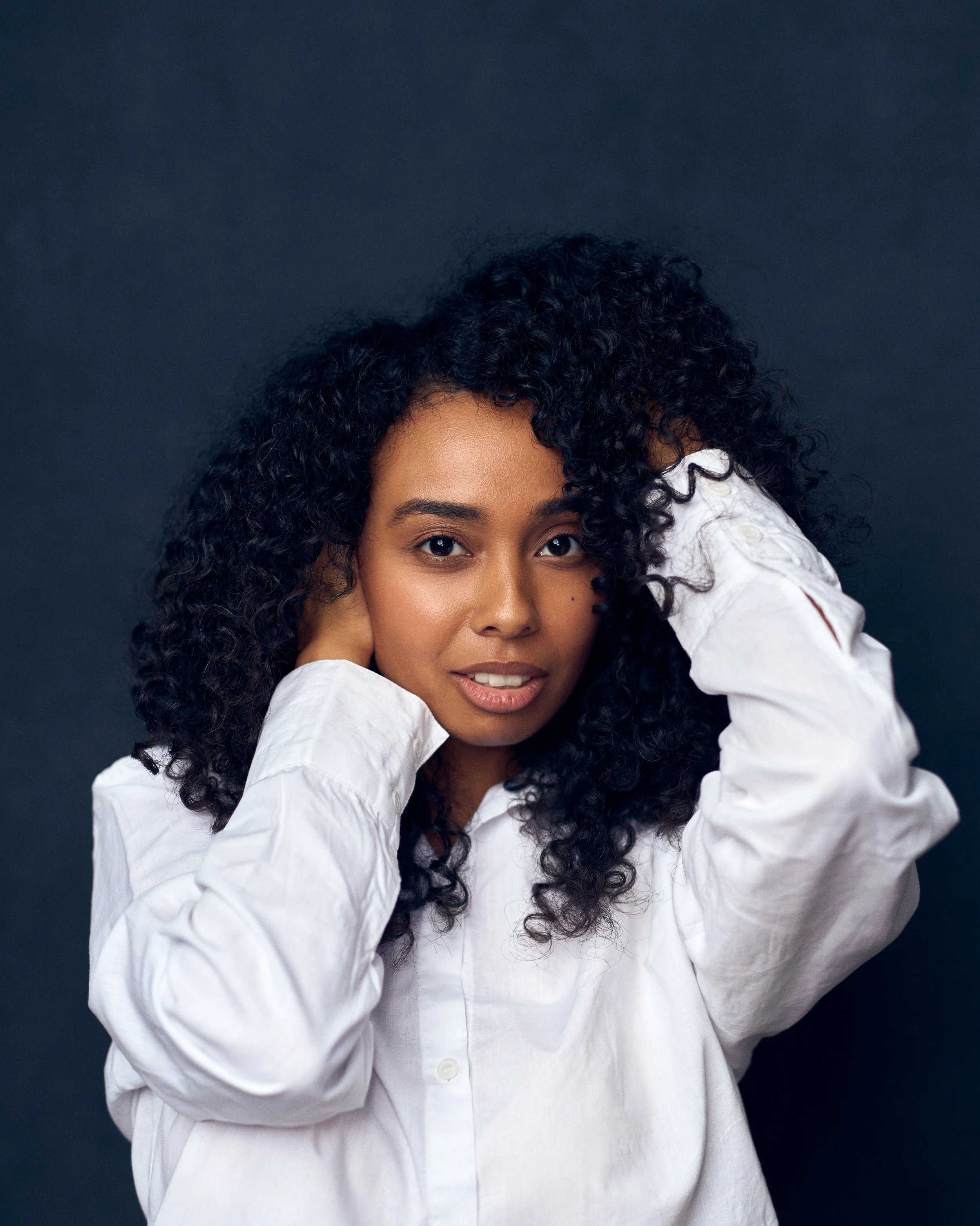 A woman with curly black hair wearing a white shirt posing against a dark background.