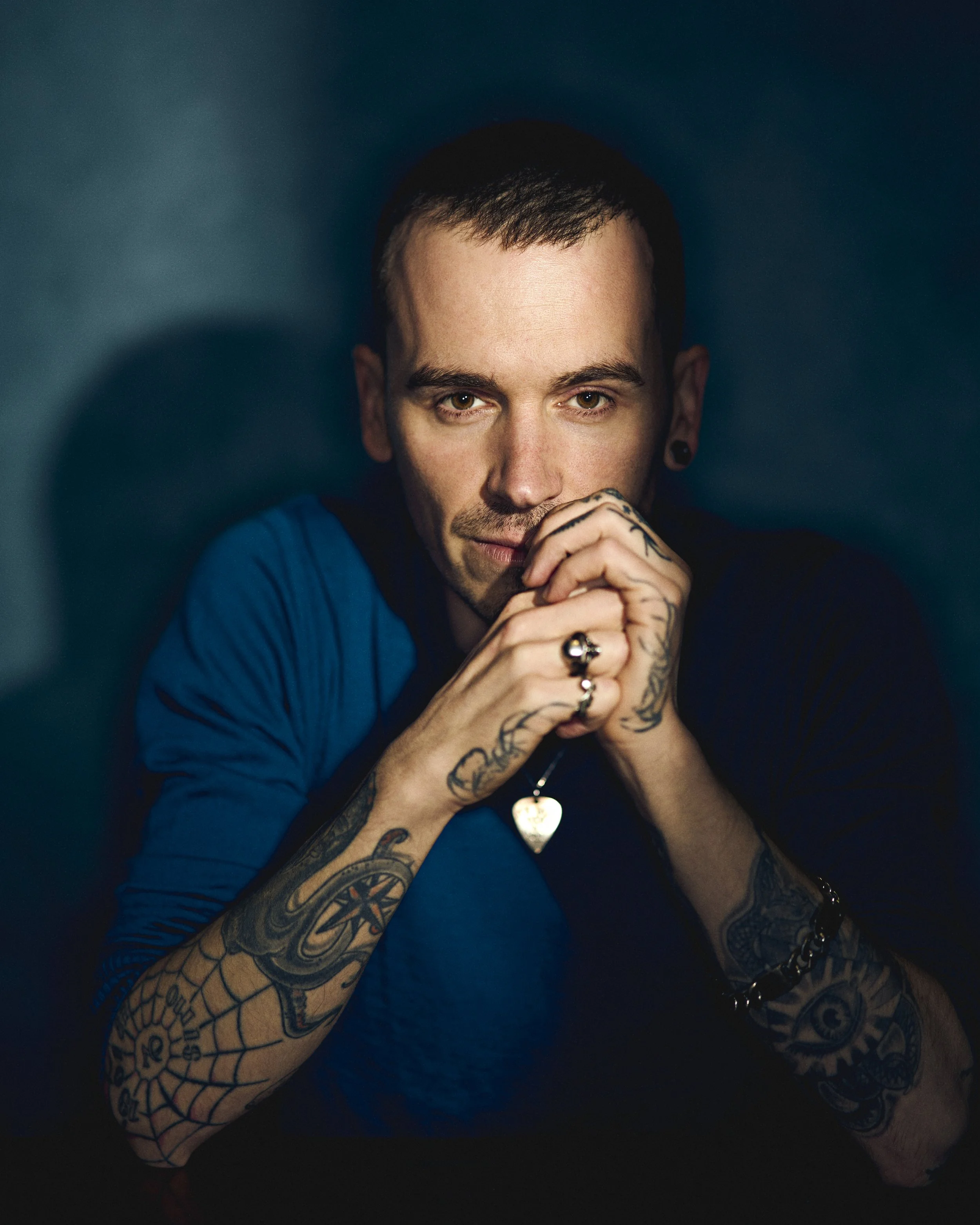 Portrait of a young man with tattoos, short dark hair, and a goatee, wearing a blue shirt and jewelry, looking at the camera with a serious expression, dark background.