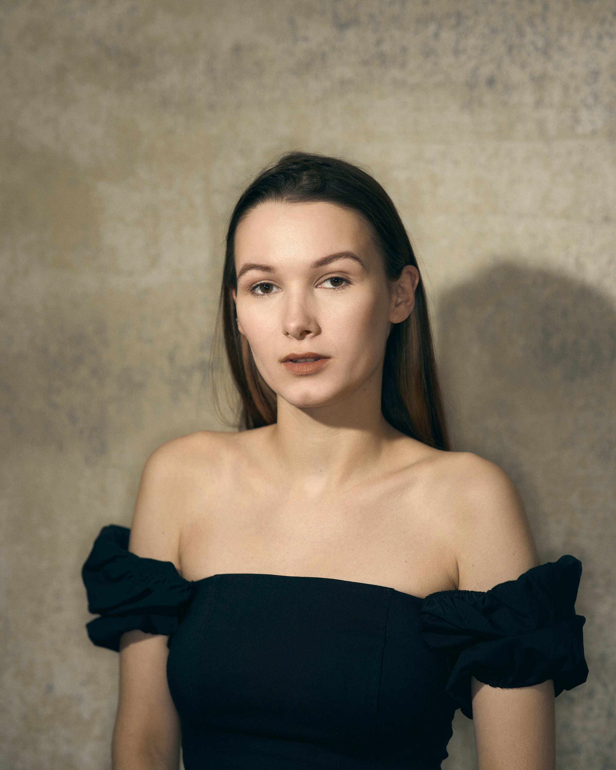 Portrait of a young woman with dark hair, wearing a black off-shoulder top, standing against a plain, textured background.