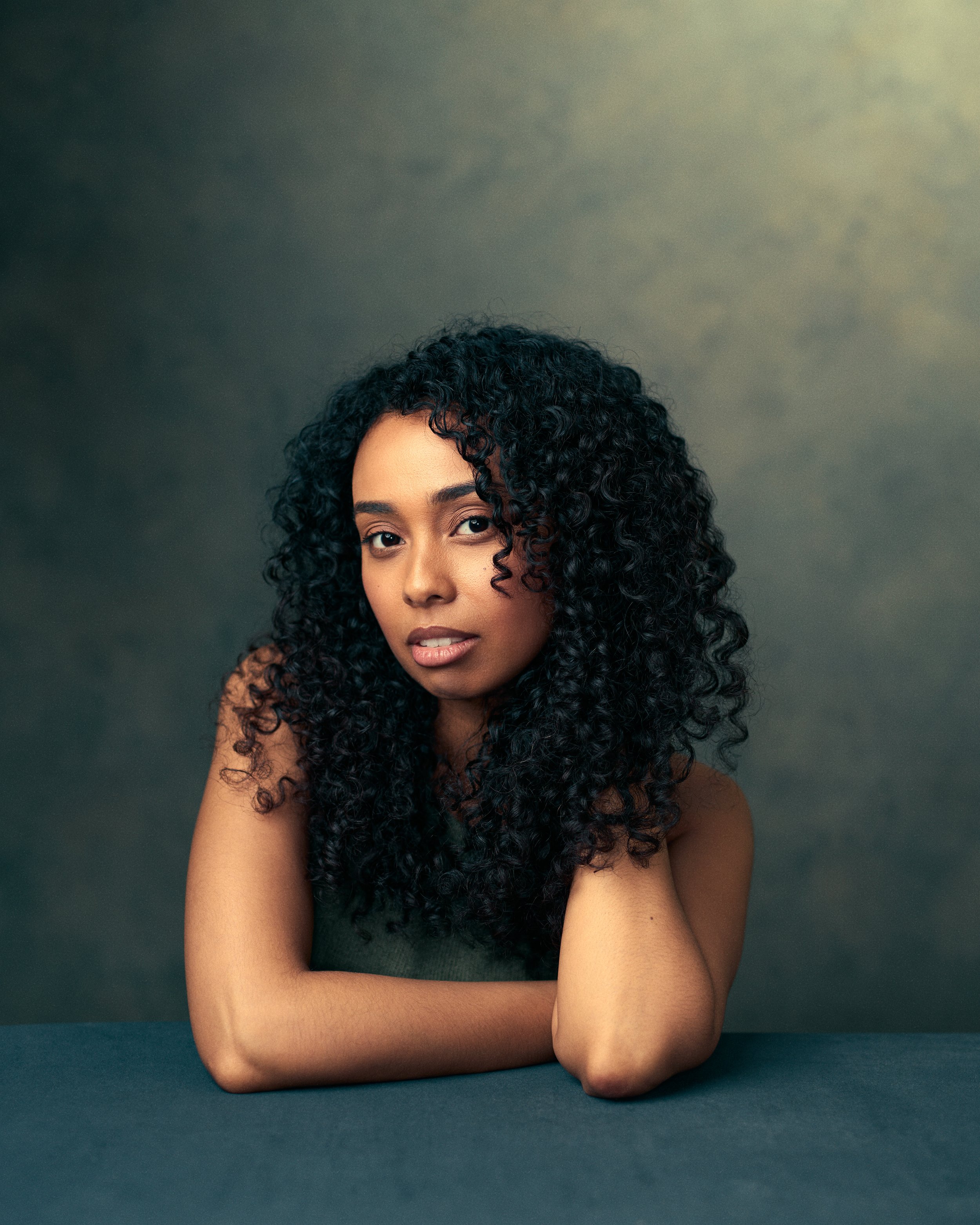 Portrait of a woman with curly black hair, resting her chin on her hand, looking at the camera, against a dark background.