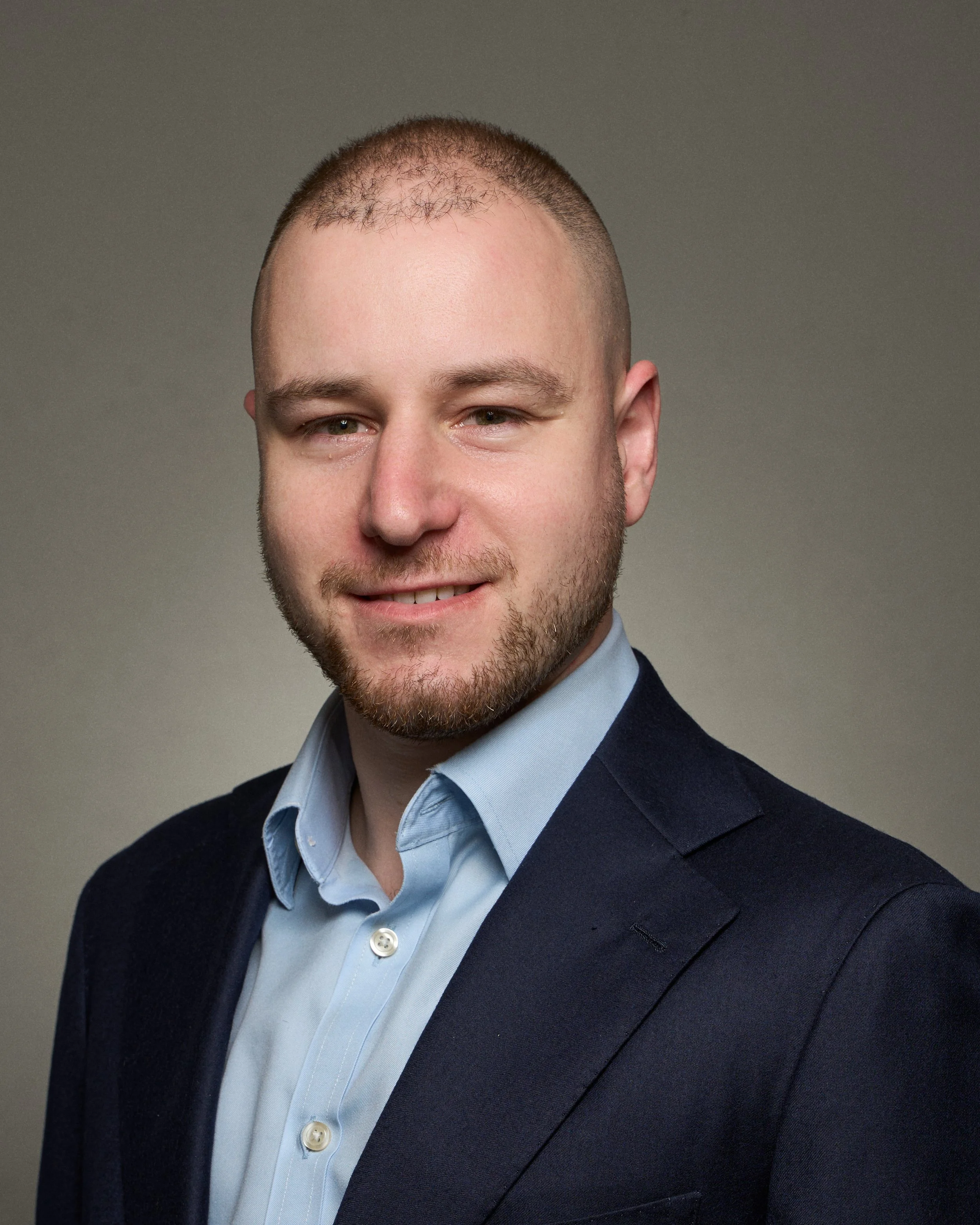 Professional headshot of a man wearing a navy blazer and light blue shirt, smiling slightly, with a neutral gray background.