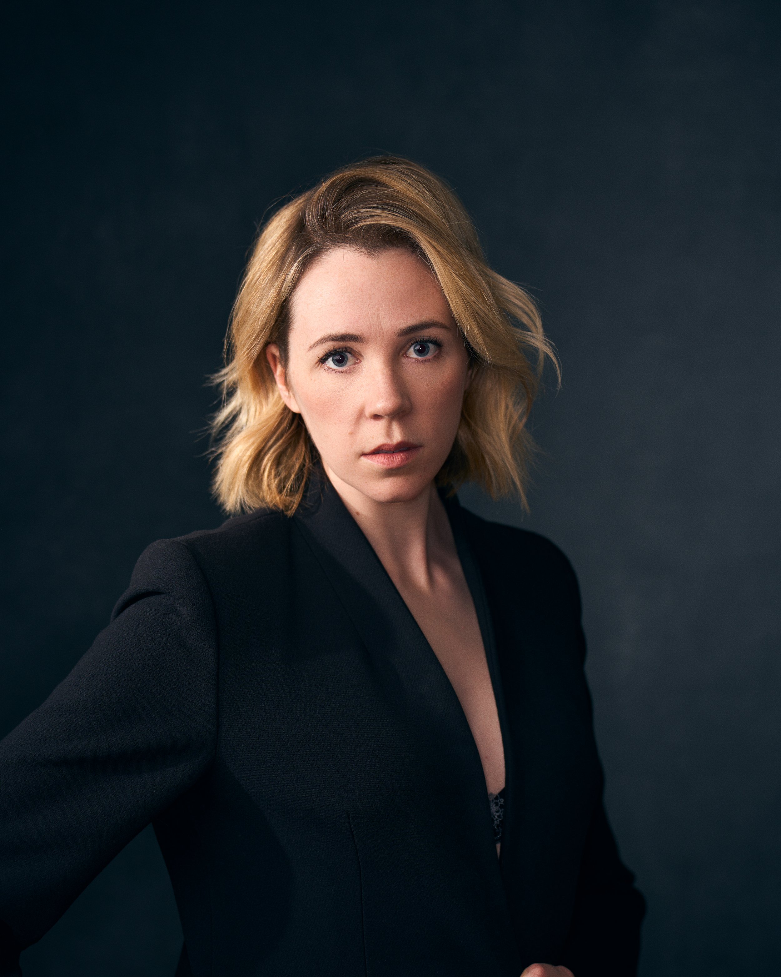 A woman with blonde hair and blue eyes wearing a black blazer, staring intensely at the camera against a dark background.