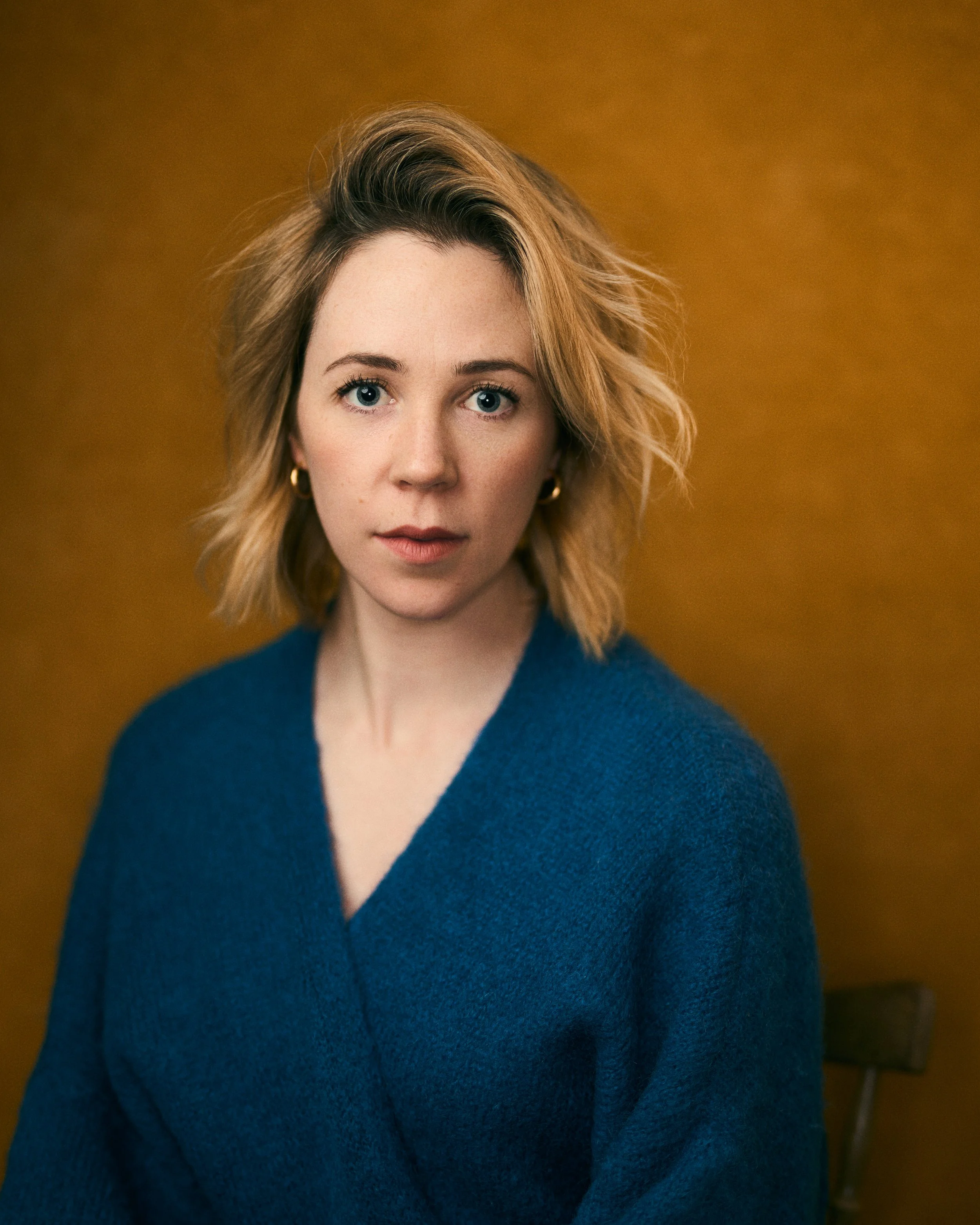 A woman with blonde, wavy hair and blue eyes wearing a blue sweater and gold hoop earrings, against a warm brown background.