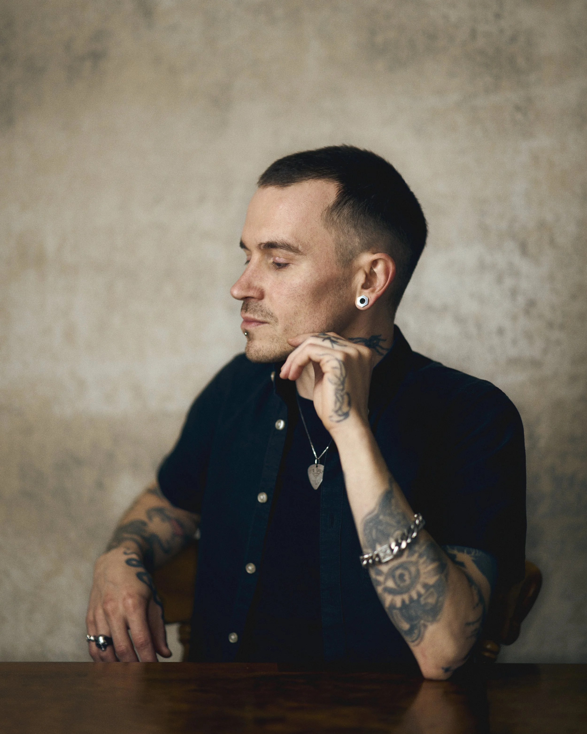 A young man with short dark hair and tattoos on his arms, wearing a black shirt and jewelry, sitting at a wooden table with a neutral beige background.