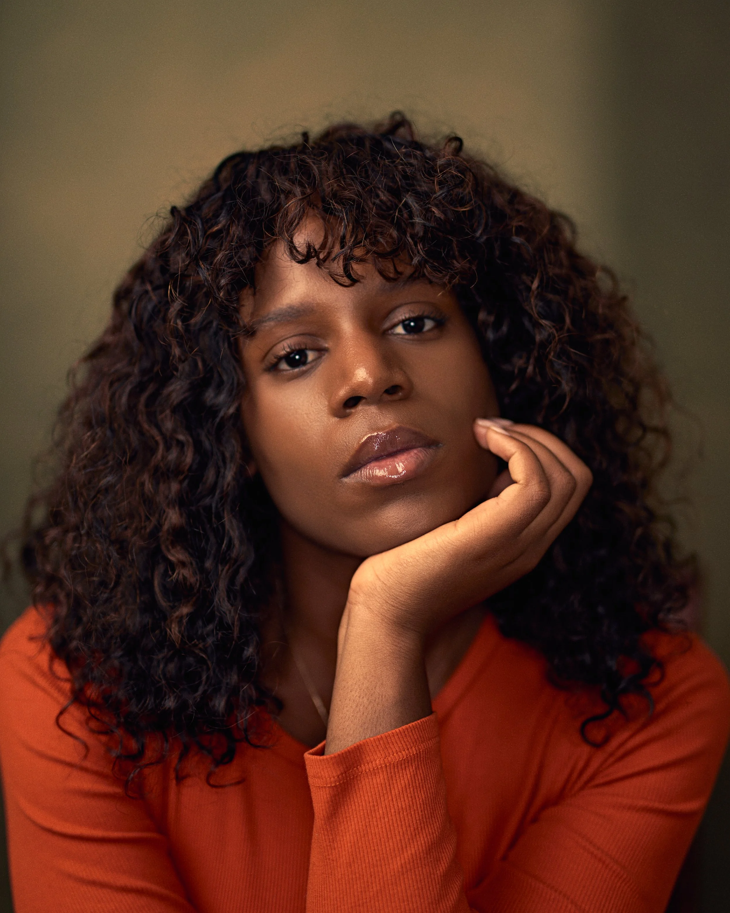A woman with curly hair resting her chin on her hand, wearing an orange top, looking at the camera with a neutral expression.