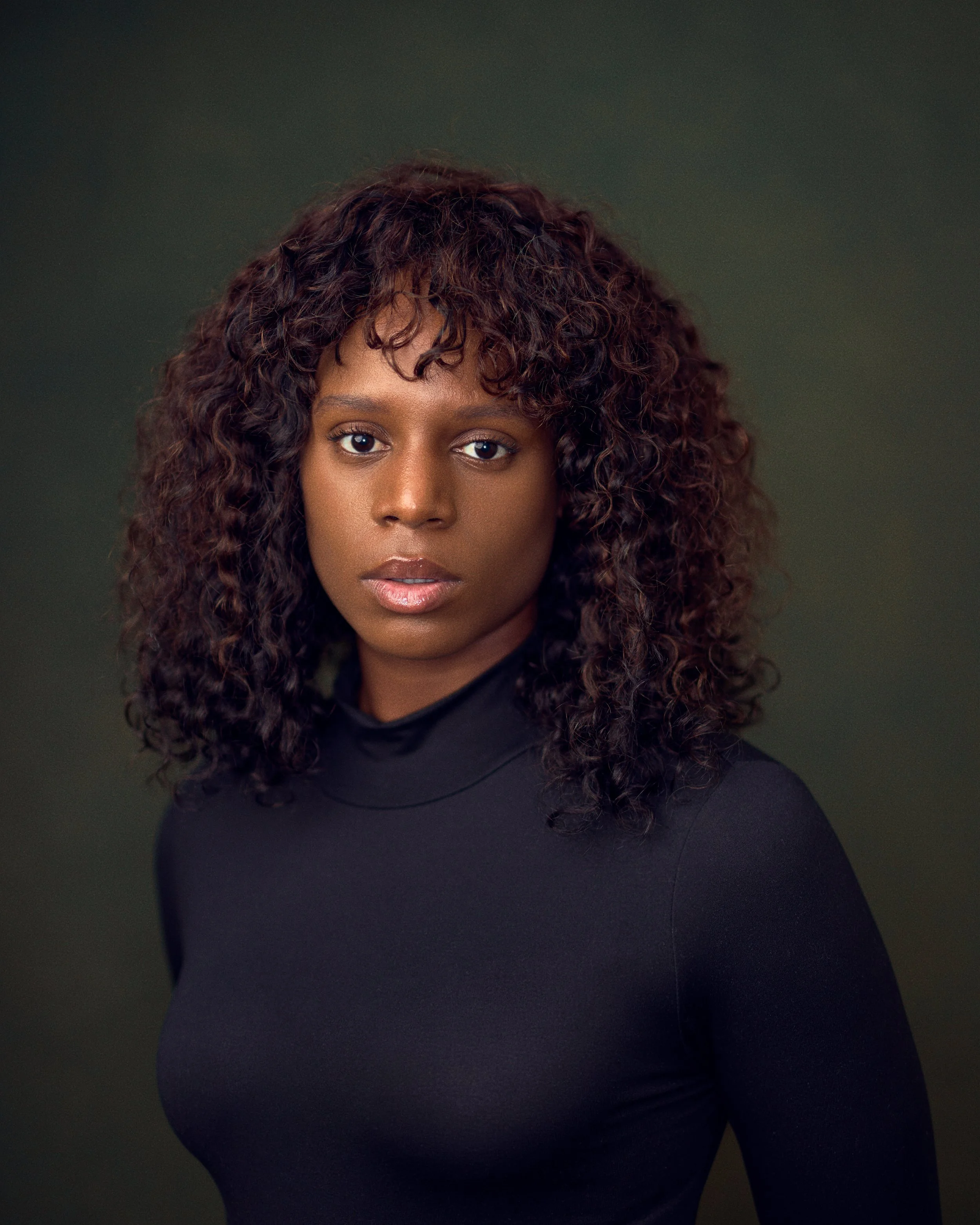 Portrait of a woman with curly hair wearing a black turtleneck against a dark background.