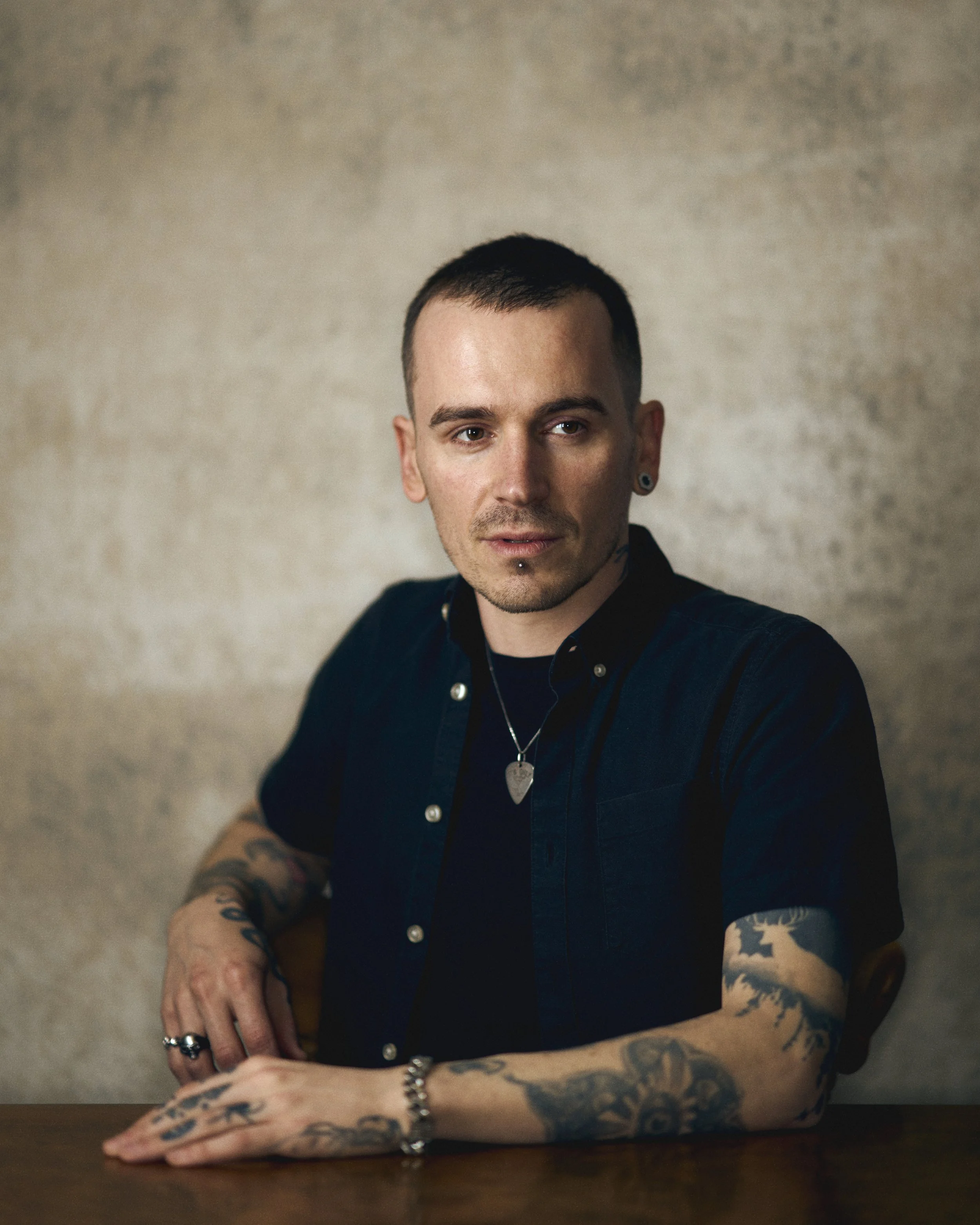 Portrait of a young man with short dark hair, tattoos on arms, wearing a black shirt and jewelry, sitting at a wooden table against a neutral background.
