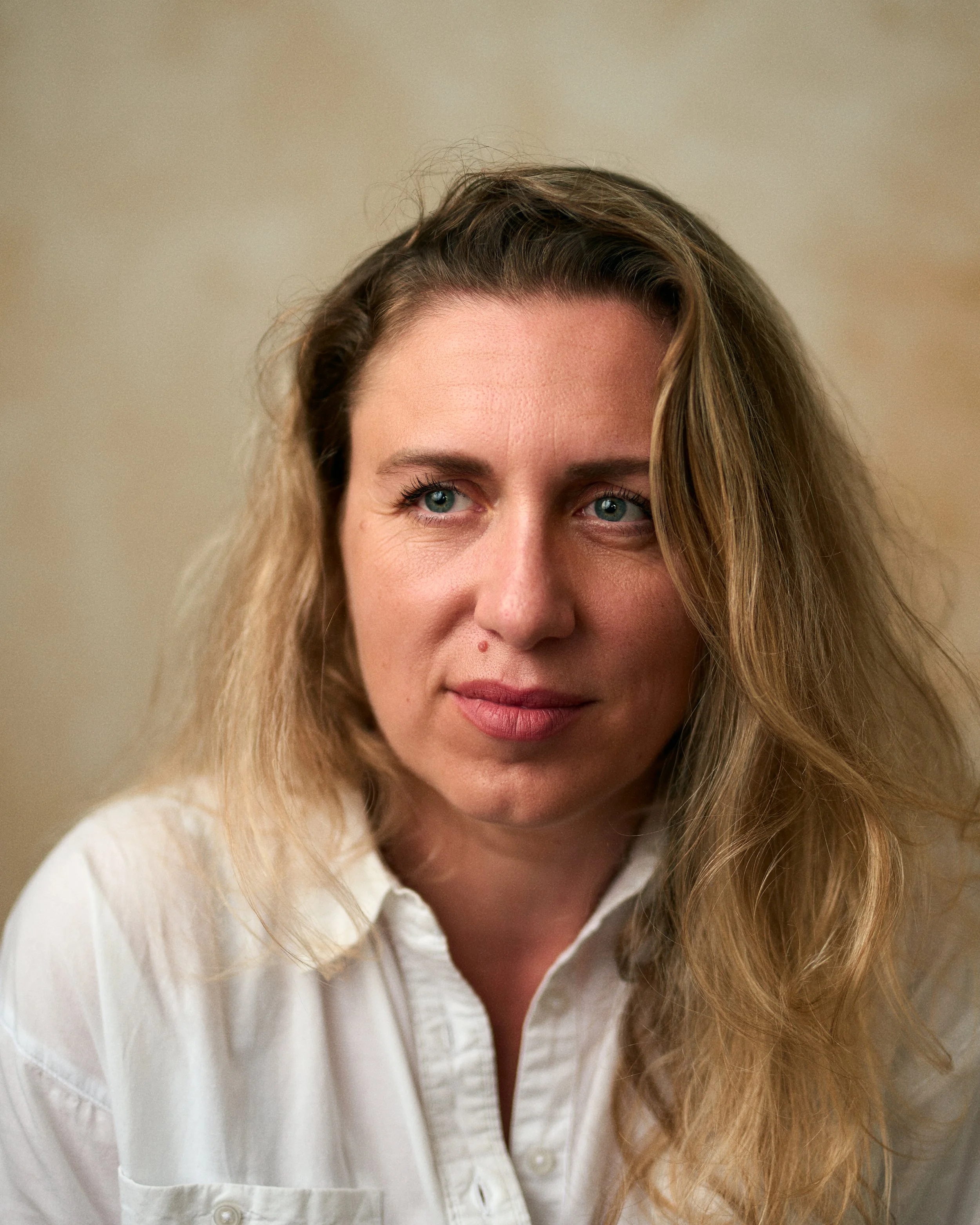 Close-up portrait of a woman with curly blonde hair, blue eyes, and light skin, wearing a white collared shirt, against a beige background.