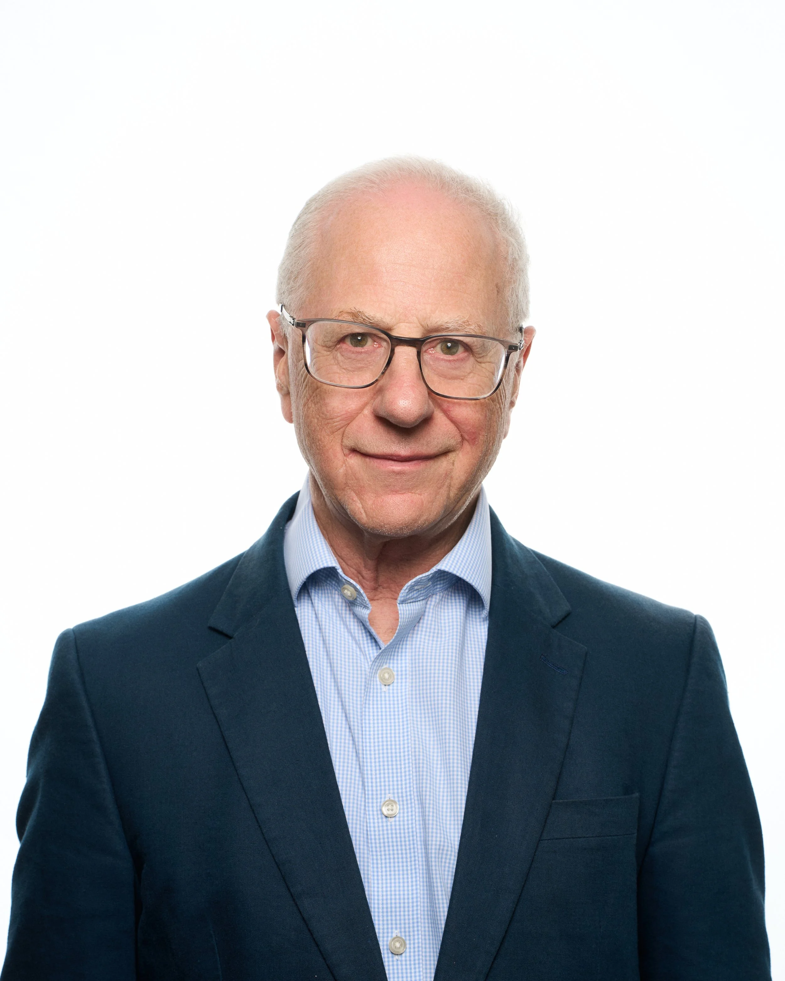 Portrait of an elderly man with glasses, wearing a navy blazer and light blue shirt, smiling against a white background.