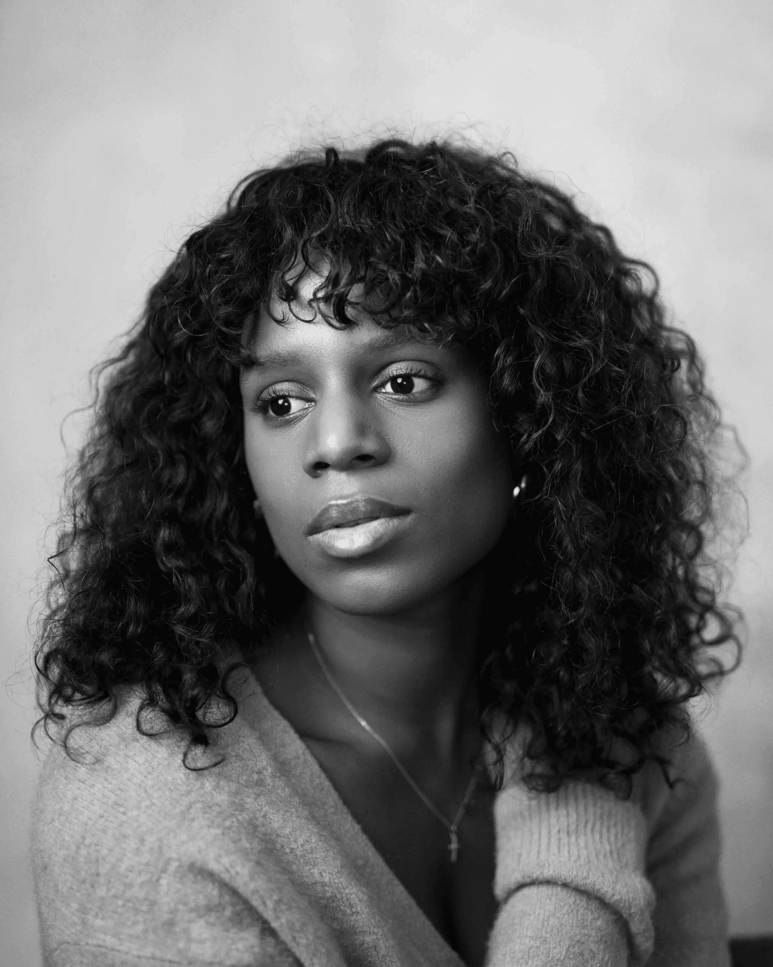 Black and white portrait of a woman with curly hair wearing a sweater, looking to the side with a thoughtful expression.