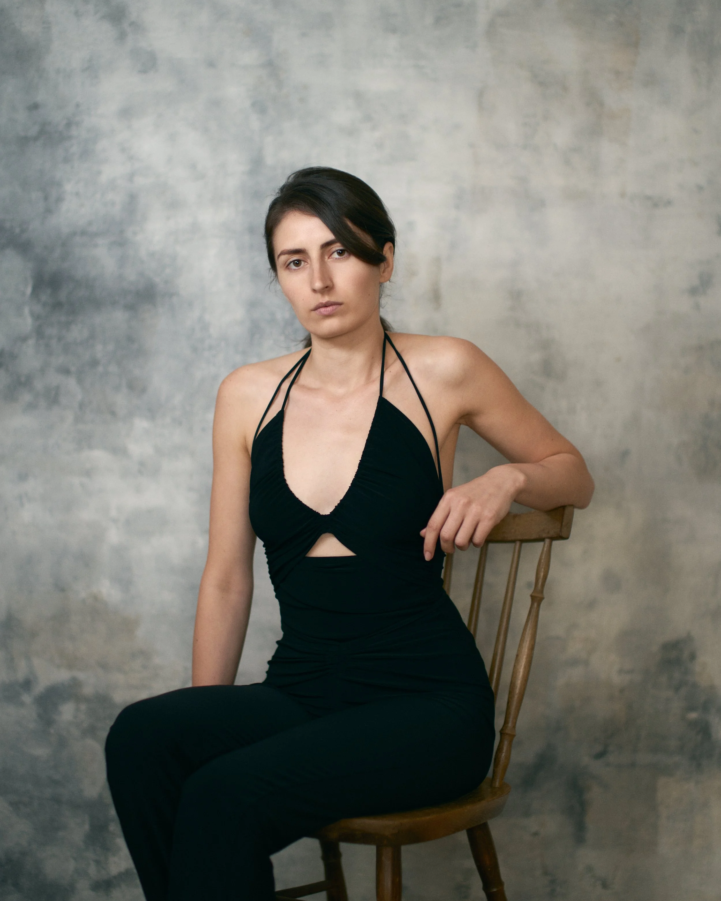 A young woman in a black dress sitting on a wooden chair against a textured gray background.