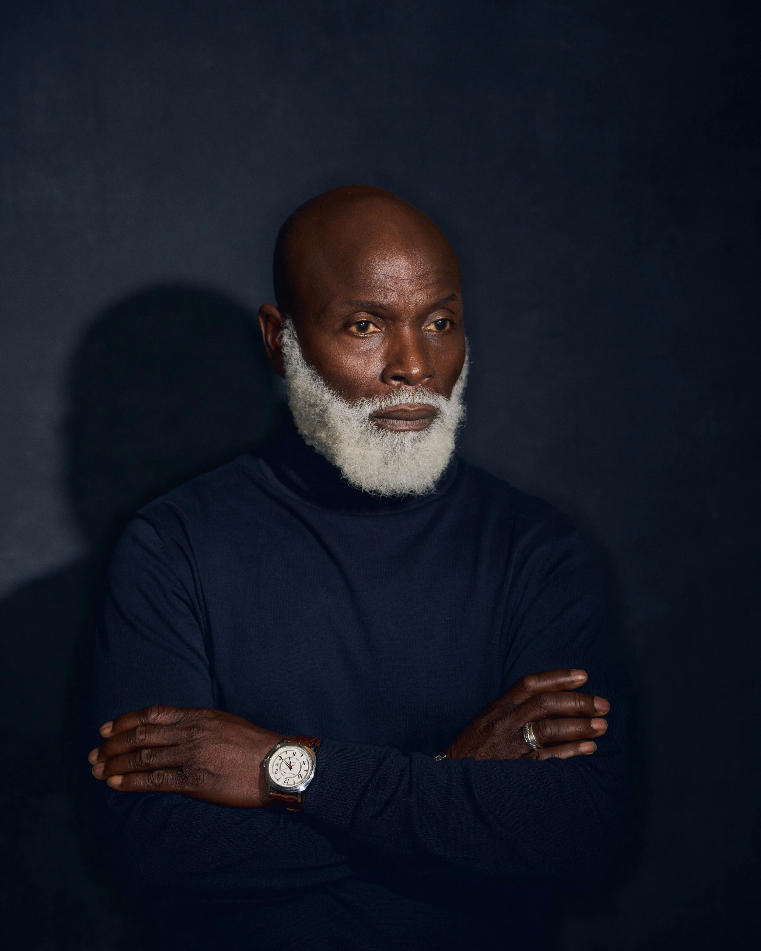 A bald man with a white beard wearing a dark blue turtleneck, crossing his arms, against a dark background with a shadow to his left.