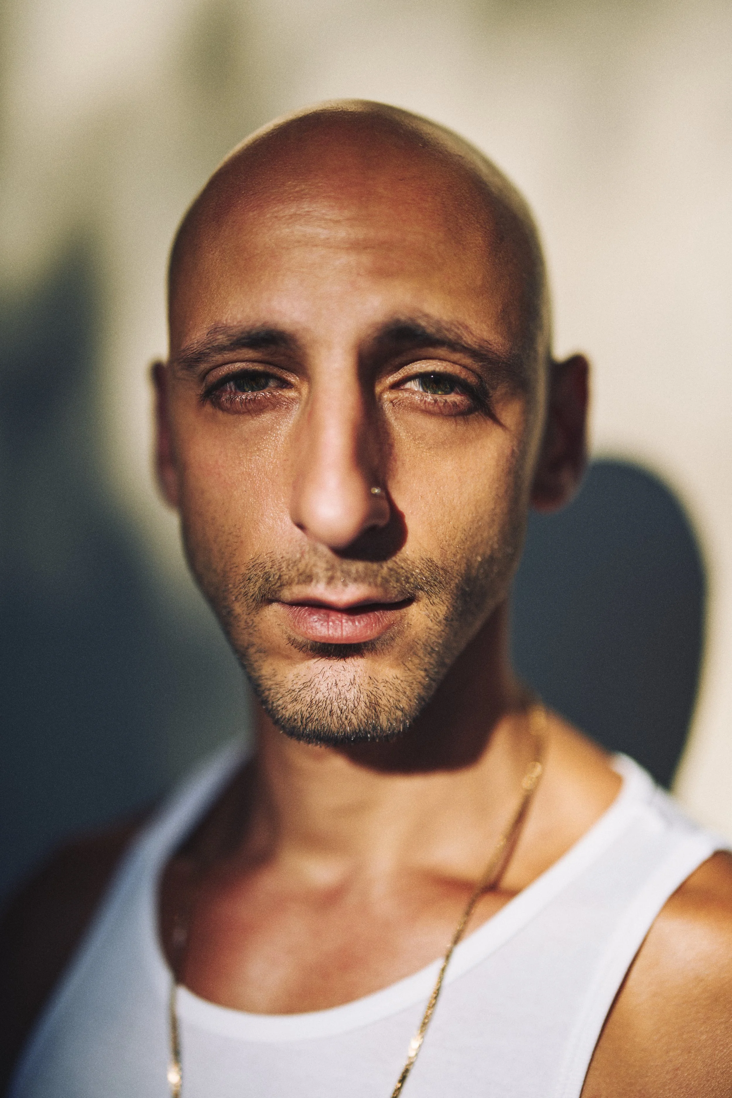 Close-up portrait of a bald man with light brown skin, wearing a white tank top and gold chain, looking at the camera with a slight smirk.