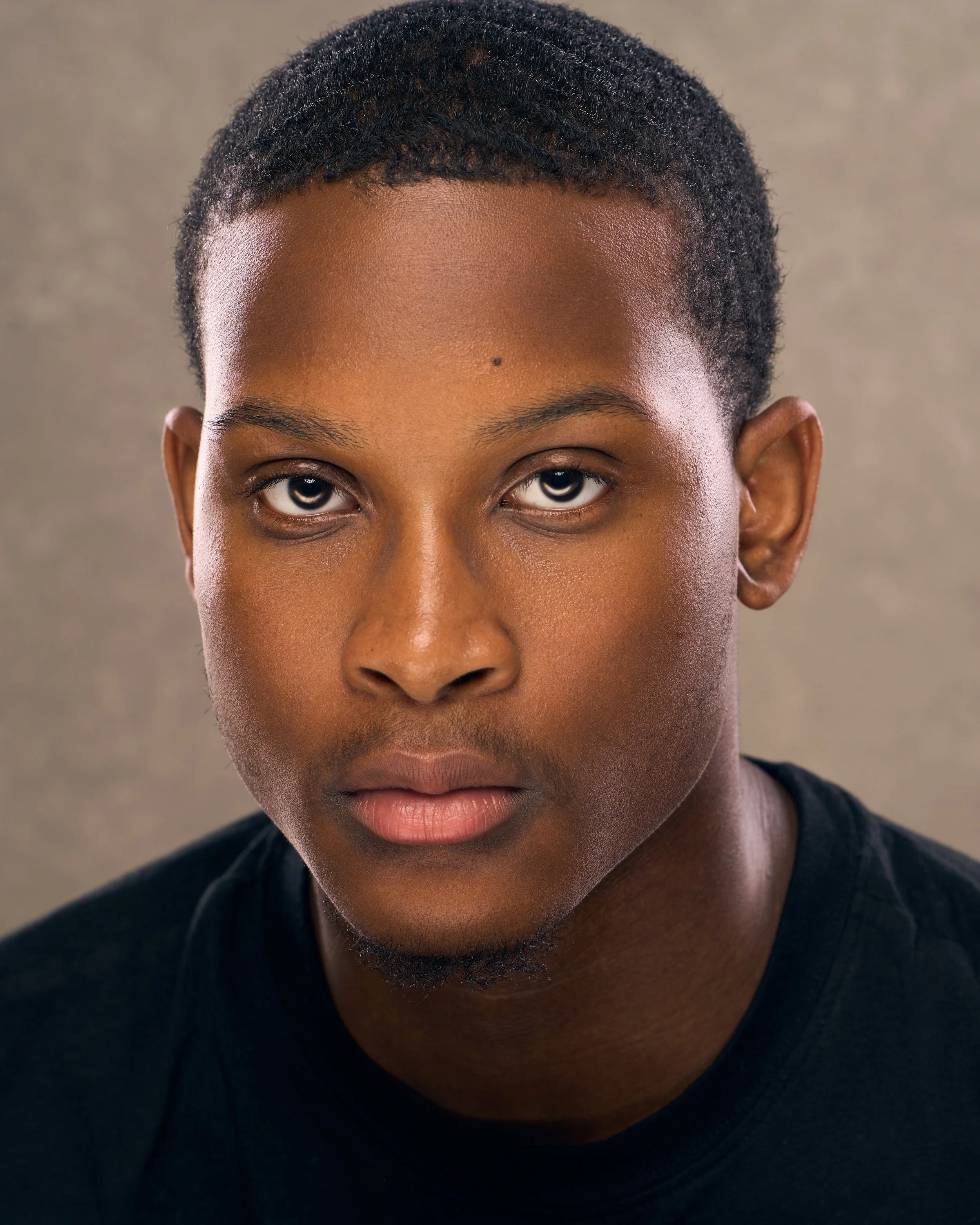 Close-up portrait of a young African American man with short hair, wearing a black shirt, against a neutral background.