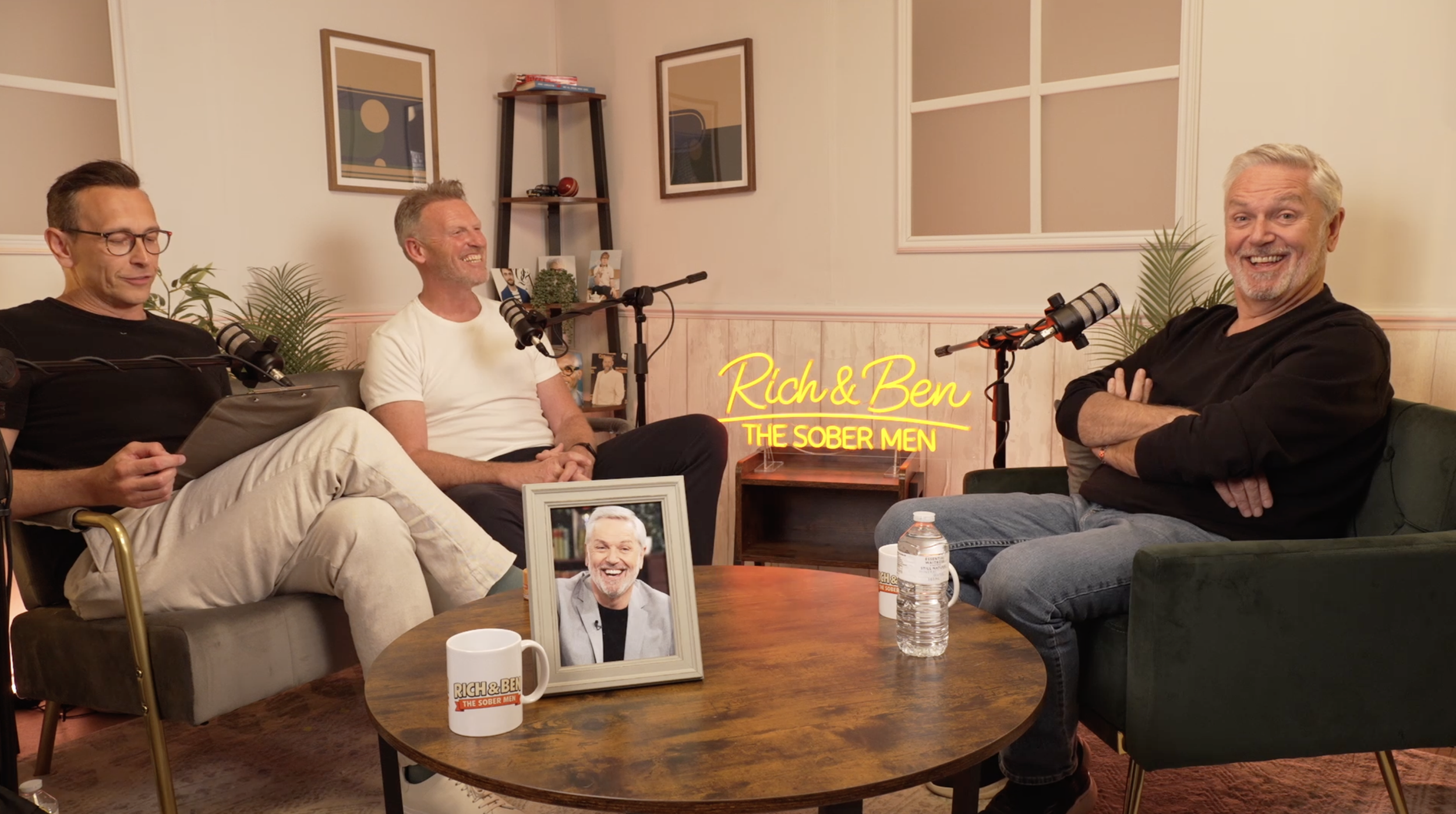 Four men sitting in a living room, participating in a podcast or interview. There are microphones, a framed photo, and a water bottle on the table. Behind them, a wooden shelf with decor, and pictures on the wall. Neon sign reads "Rich & Ben The Sober Men."