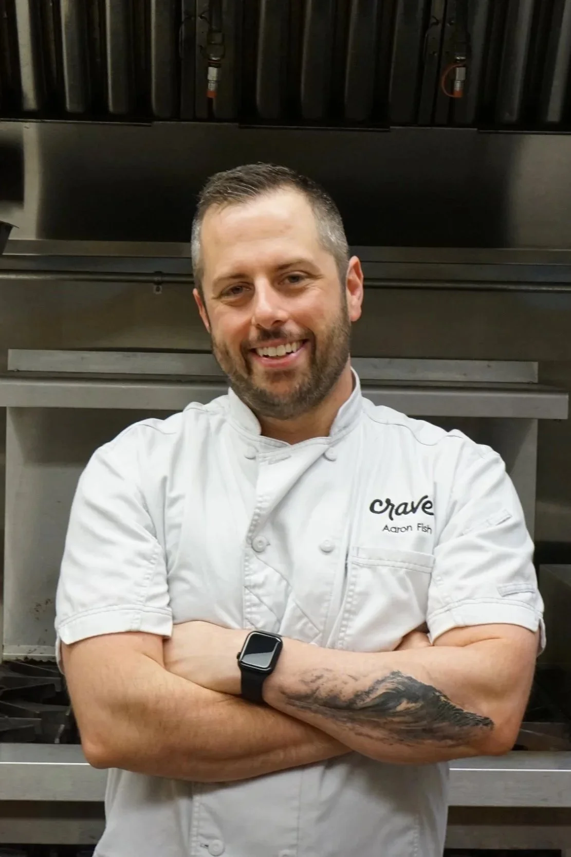 A smiling male chef in a white uniform with "Crave" and "Aaron Fish" embroidered on it, standing with arms crossed in a professional kitchen.