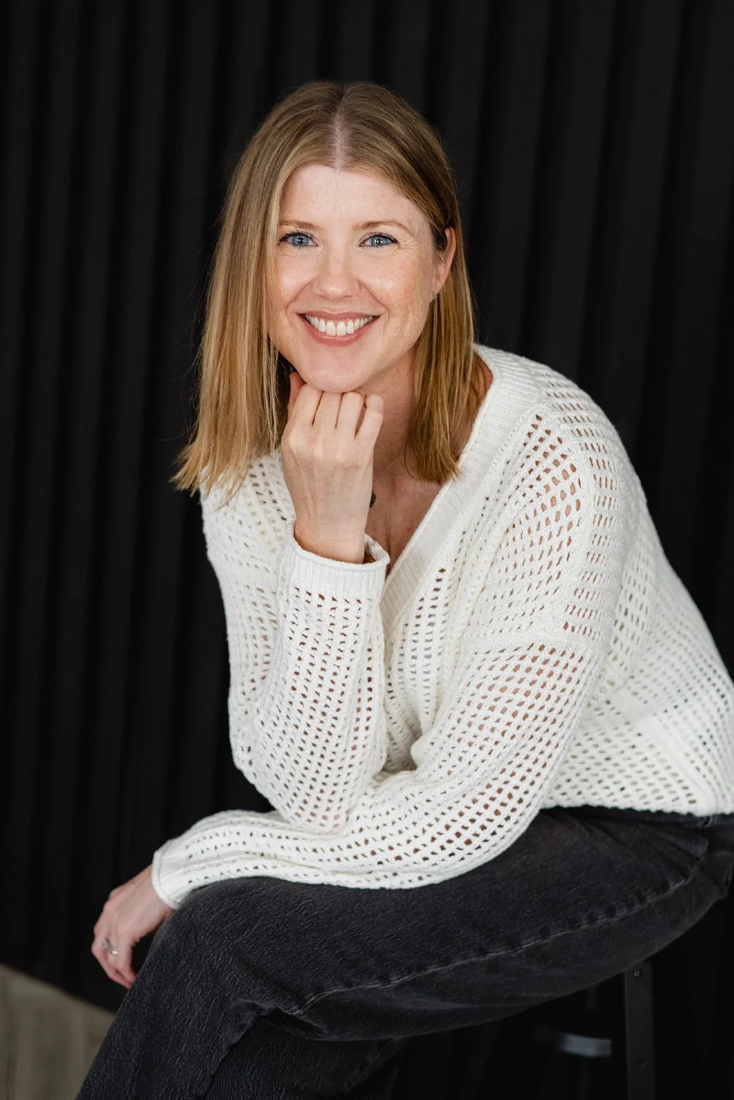 A woman with shoulder-length red hair, smiling, wearing a white knit sweater and black pants, sitting against a dark background.
