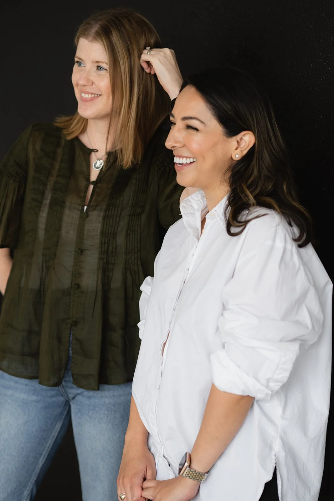 Two women laughing and smiling against a black background, one with red hair and the other with dark brown hair, dressed casually.