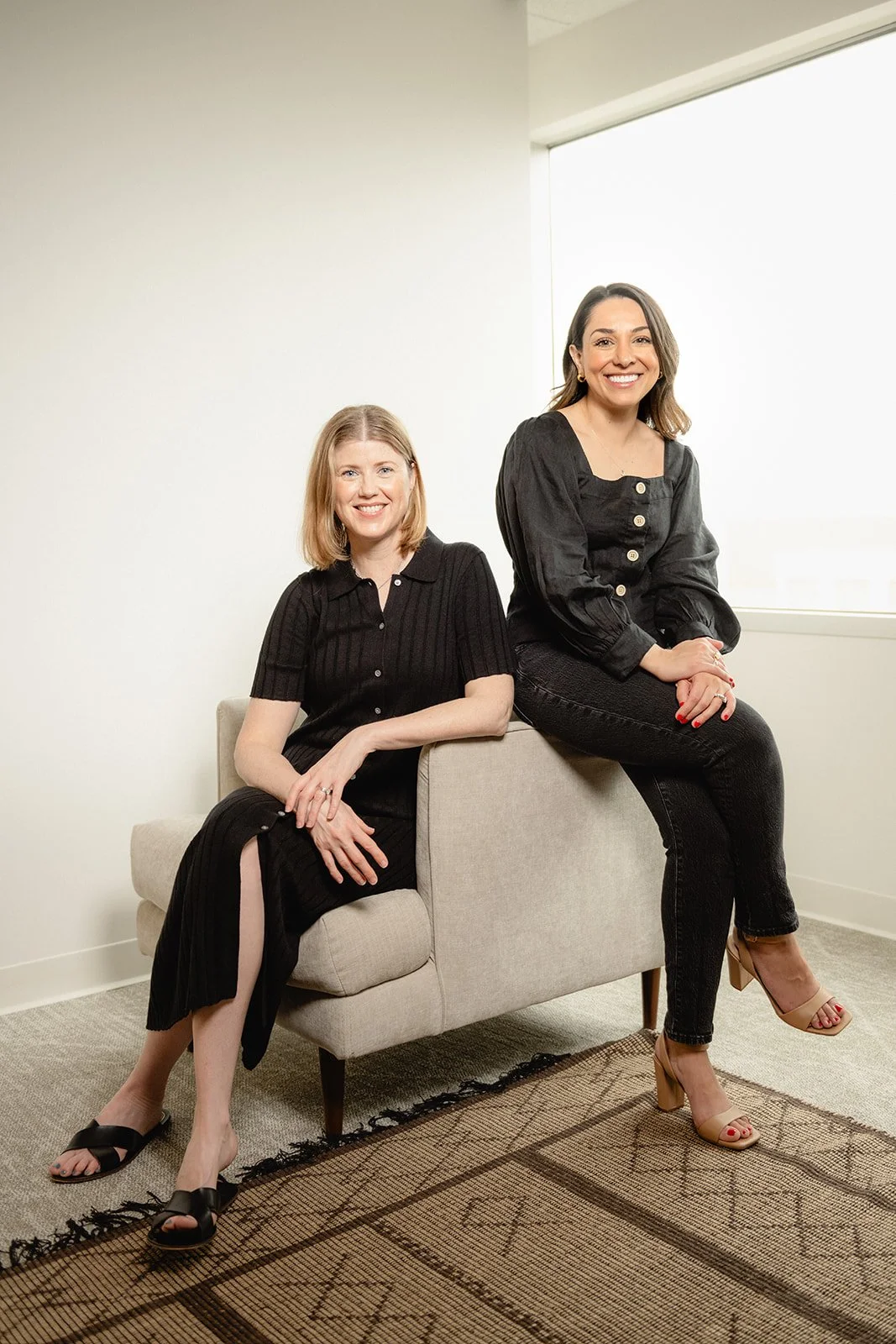 Two women in black outfits sitting on a beige sofa in front of a large window in a bright room.