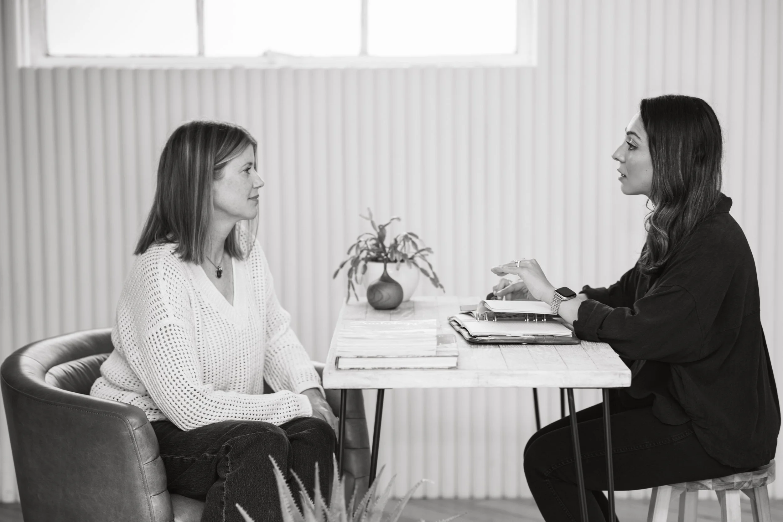 A woman in a white sweater and dark pants sitting across from a woman in a black shirt, having a conversation at a table with notebooks and a small potted plant inside a bright room.