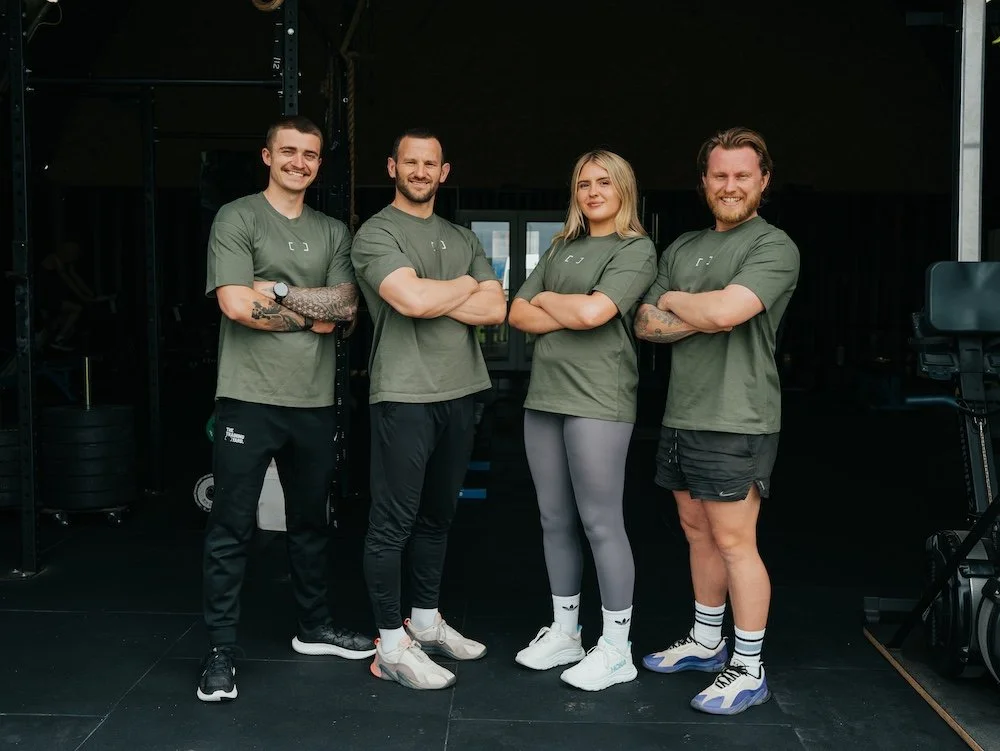 Four fitness trainers standing with arms crossed inside a gym, wearing matching green t-shirts, black or gray athletic pants or shorts, and sneakers.