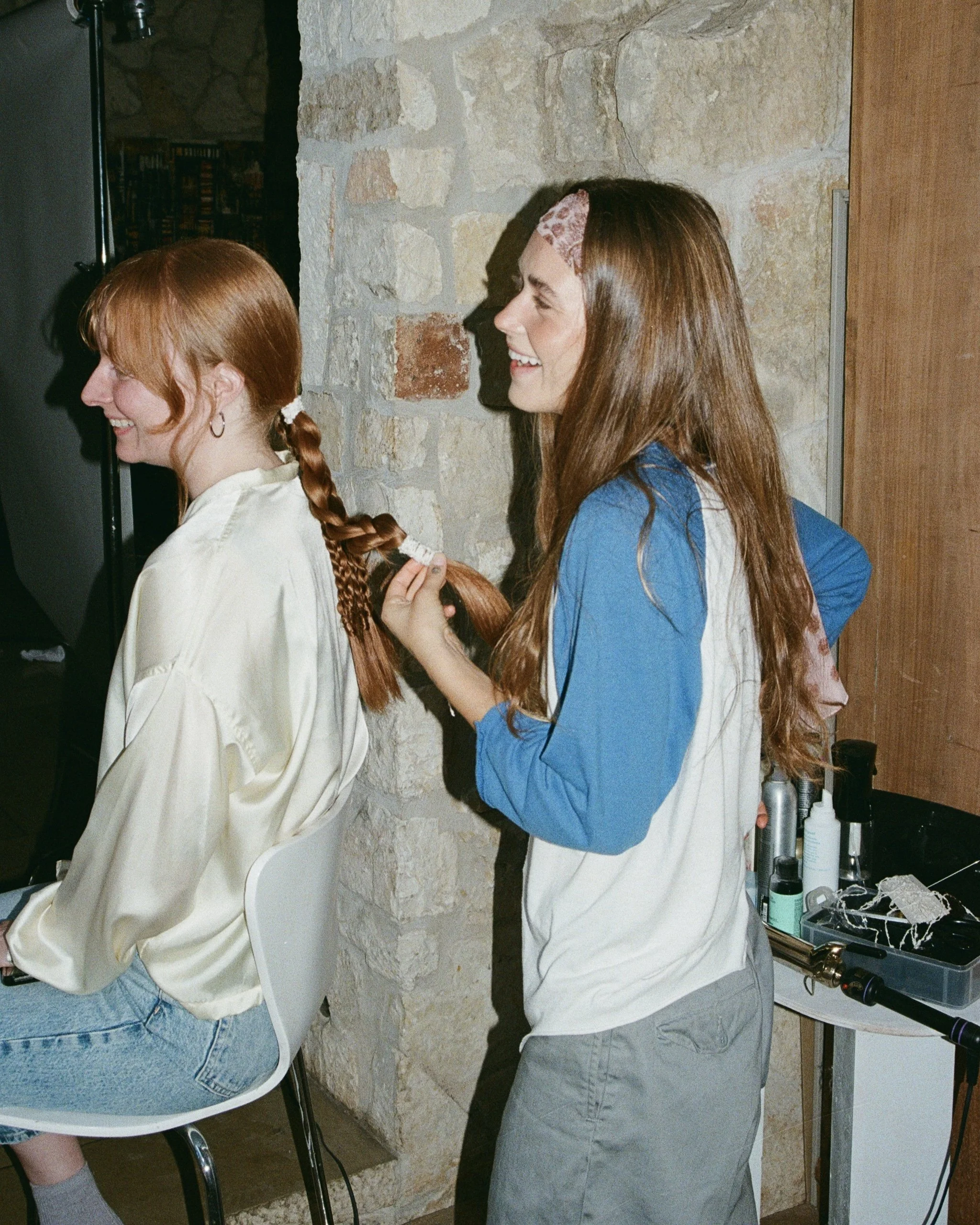 A woman with long brown hair and a pink headband is styling a red-haired woman's braid in a salon or studio with a stone wall. Both women are smiling and laughing.