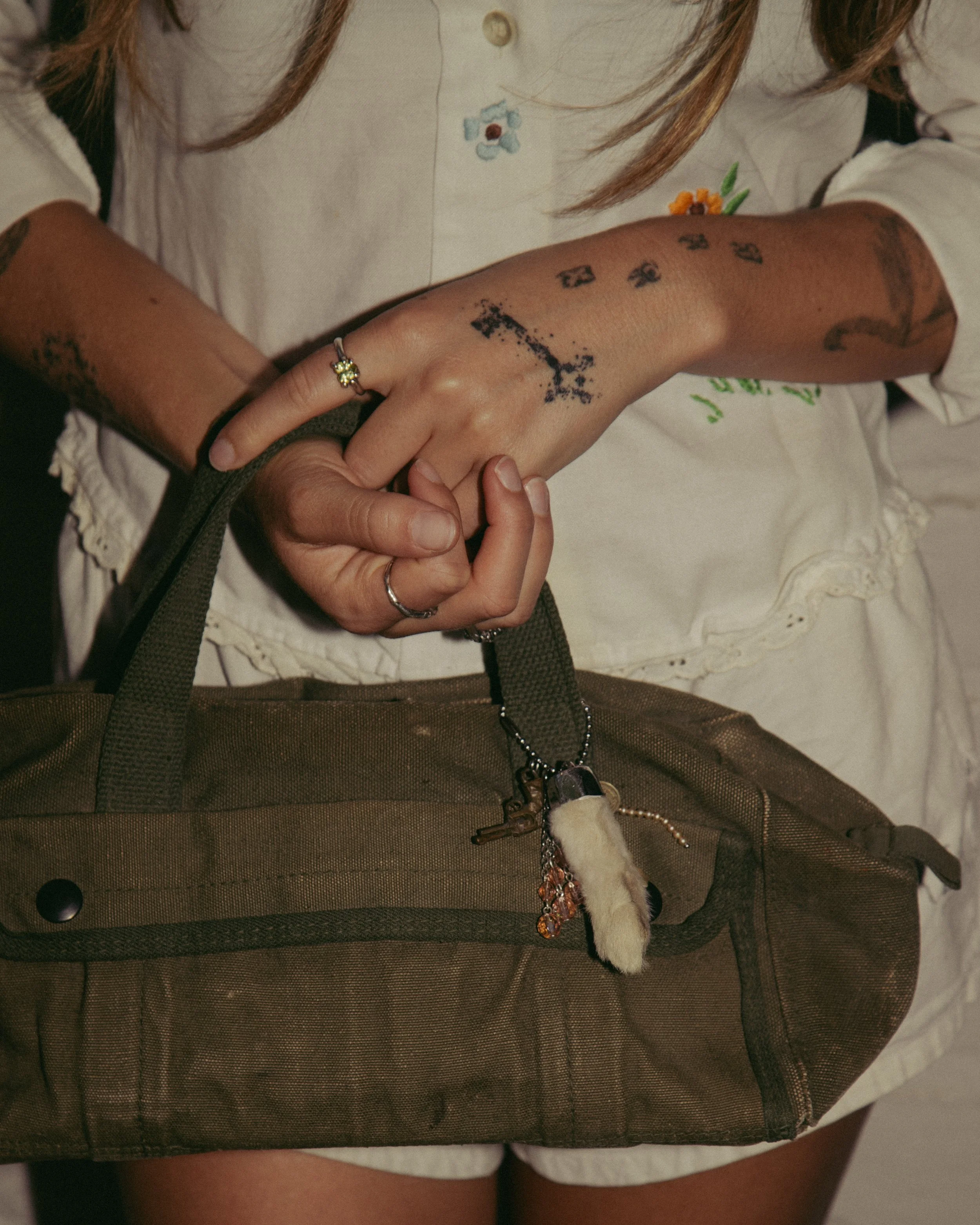Person holding a brown bag with keychain accessories, displaying tattoos on their right arm and hand, wearing multiple rings and a flower-shaped ring on their left hand.